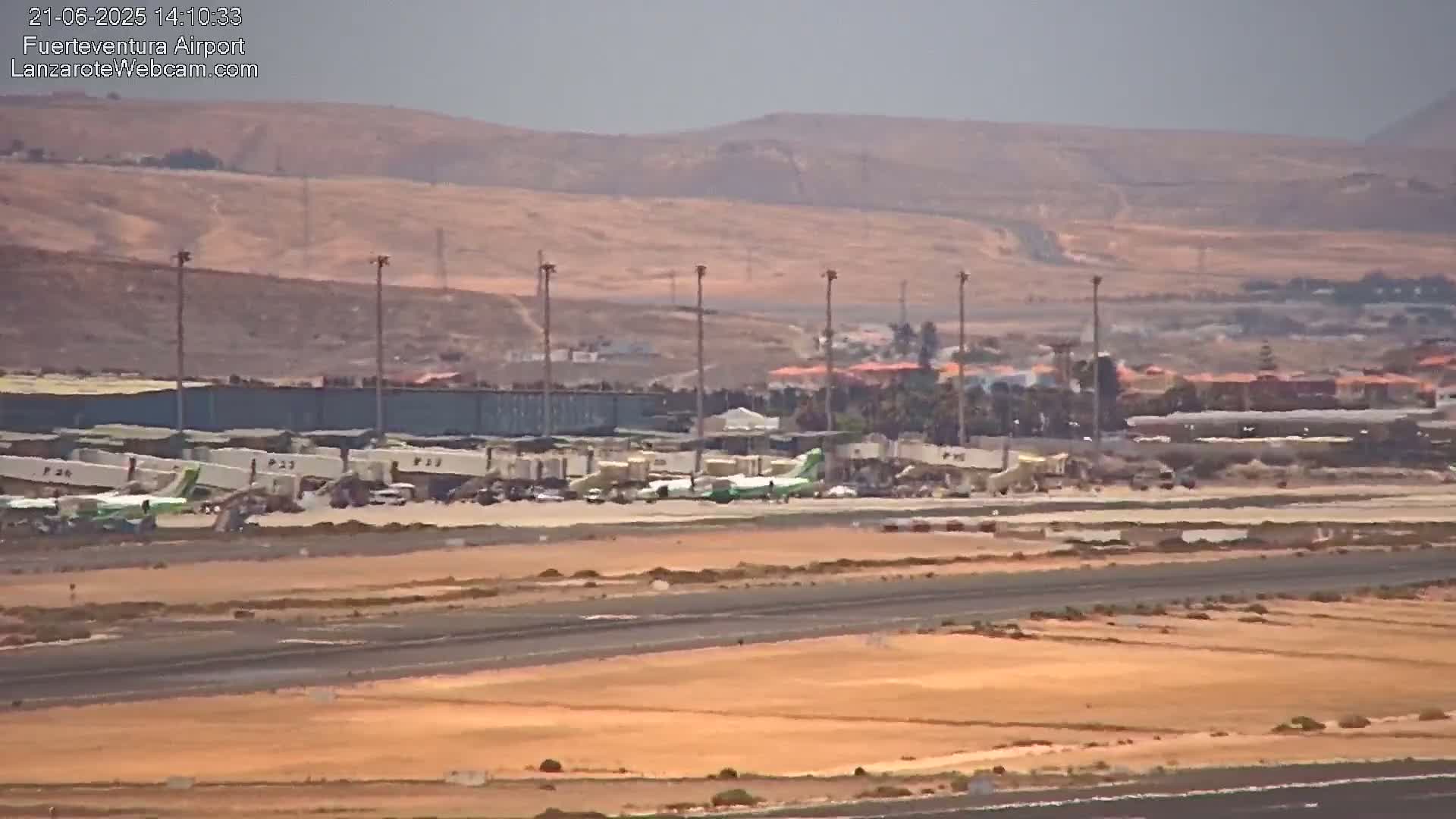 Several airplanes are parked at gates at an airport on a hazy day, with a runway and dry, light brown landscape visible in the foreground.