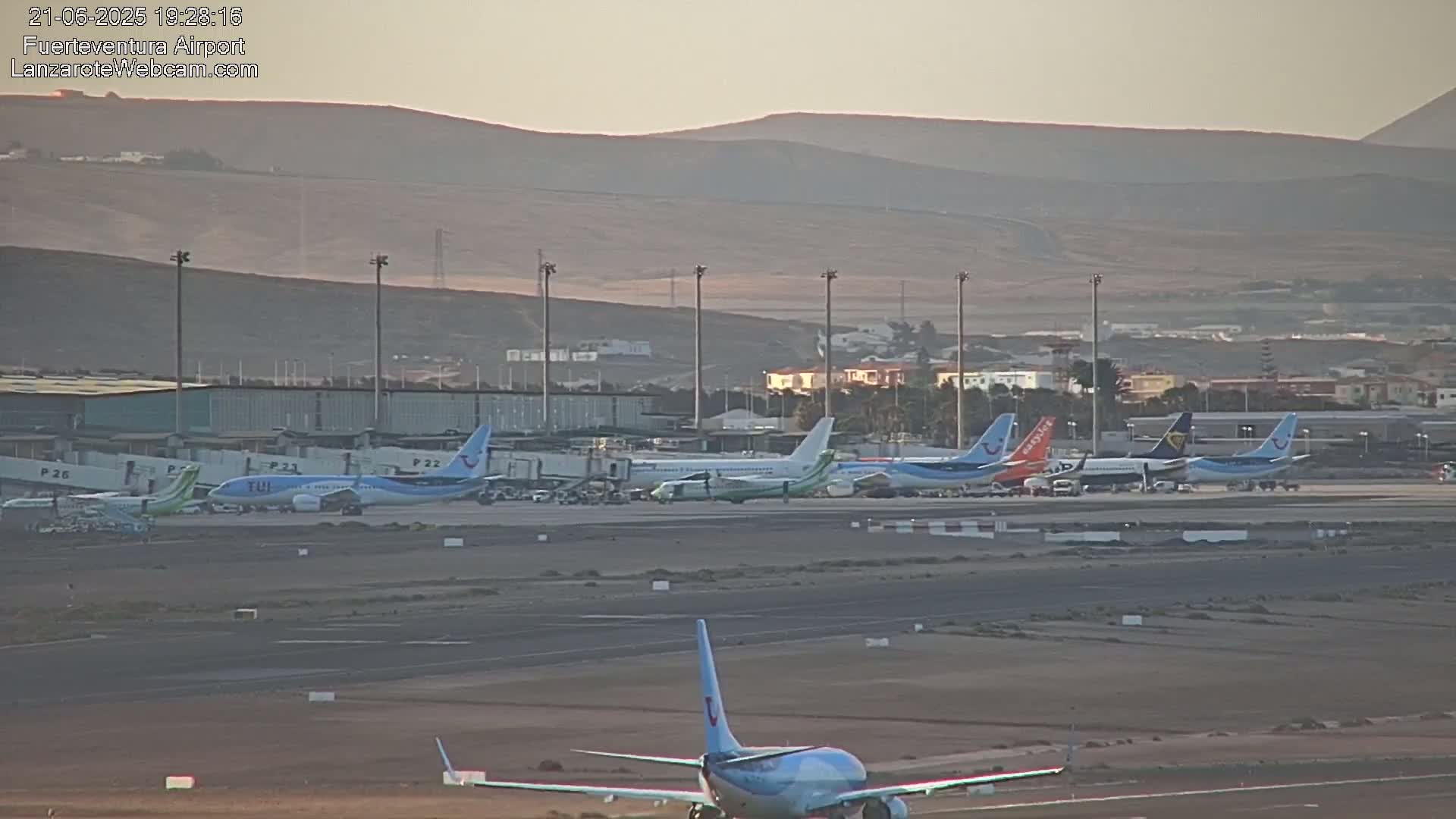 Several airplanes are parked at an airport tarmac under hazy, sunny conditions with distant mountains in the background.