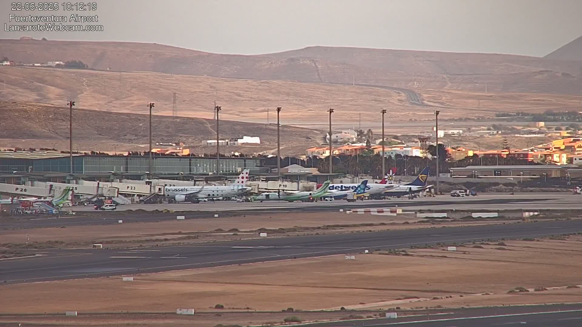 Several airplanes are parked at gates at an airport, with a brownish-tan landscape visible in the background under a clear sky.