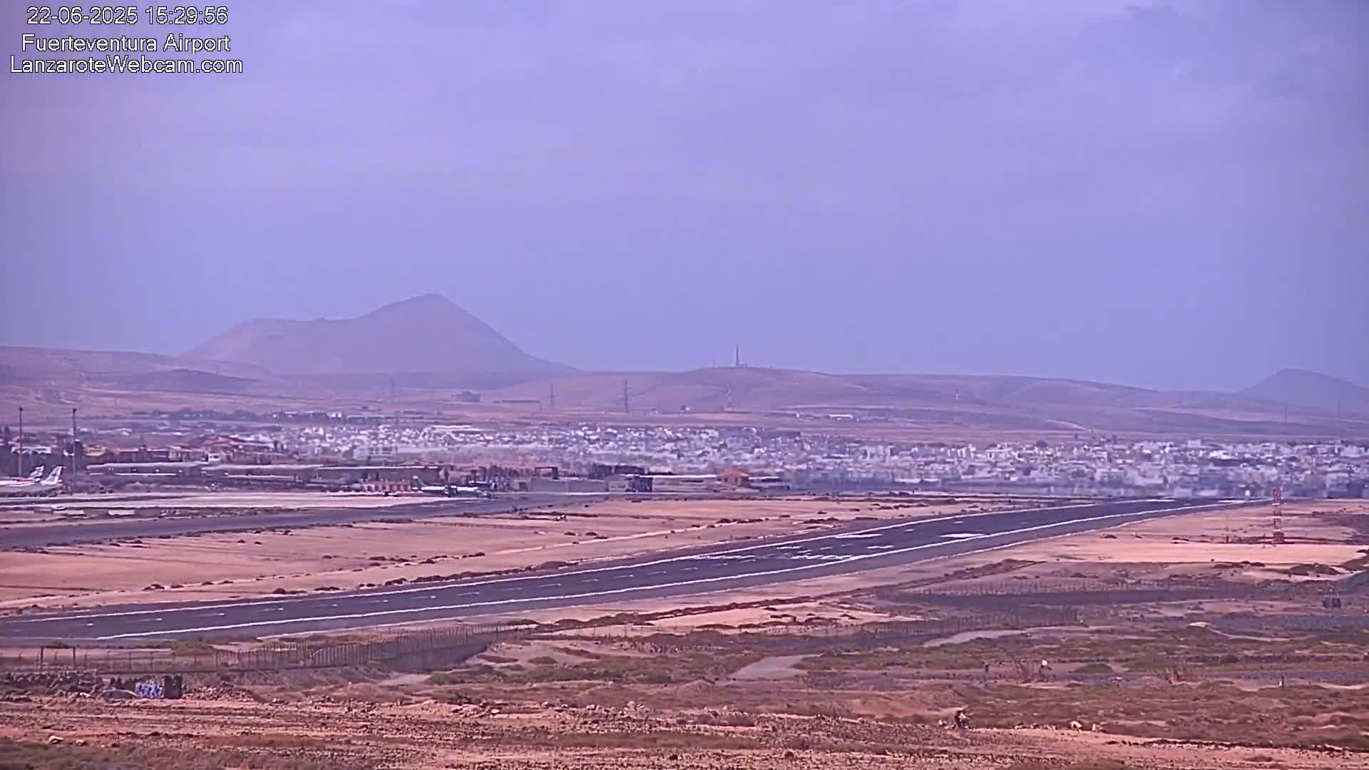A long runway stretches across a flat, desert-like landscape toward a town and distant mountains under a hazy sky.