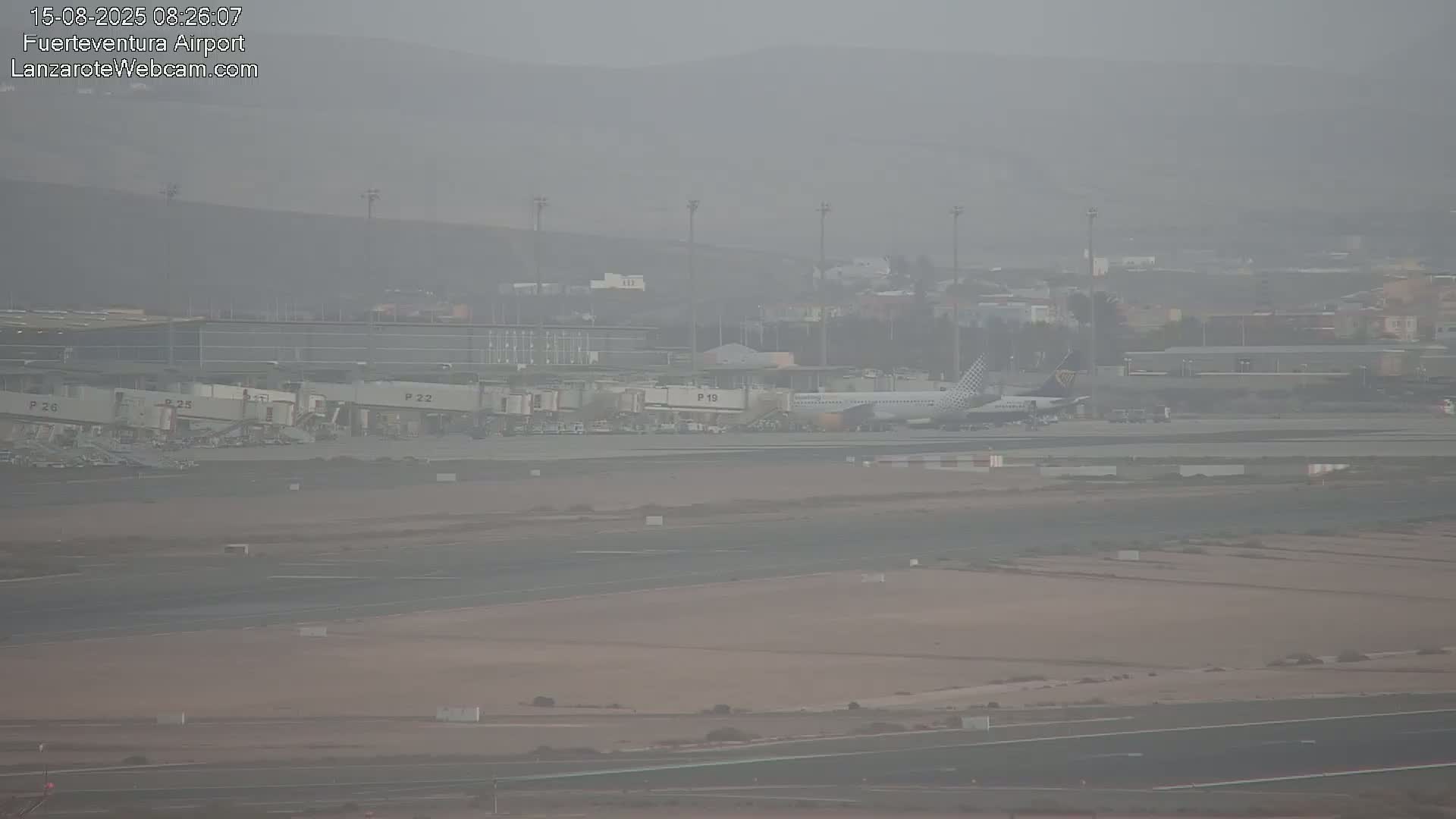 A hazy view of an airport tarmac shows a passenger plane at a gate, with several other gates and airport buildings visible in the background.