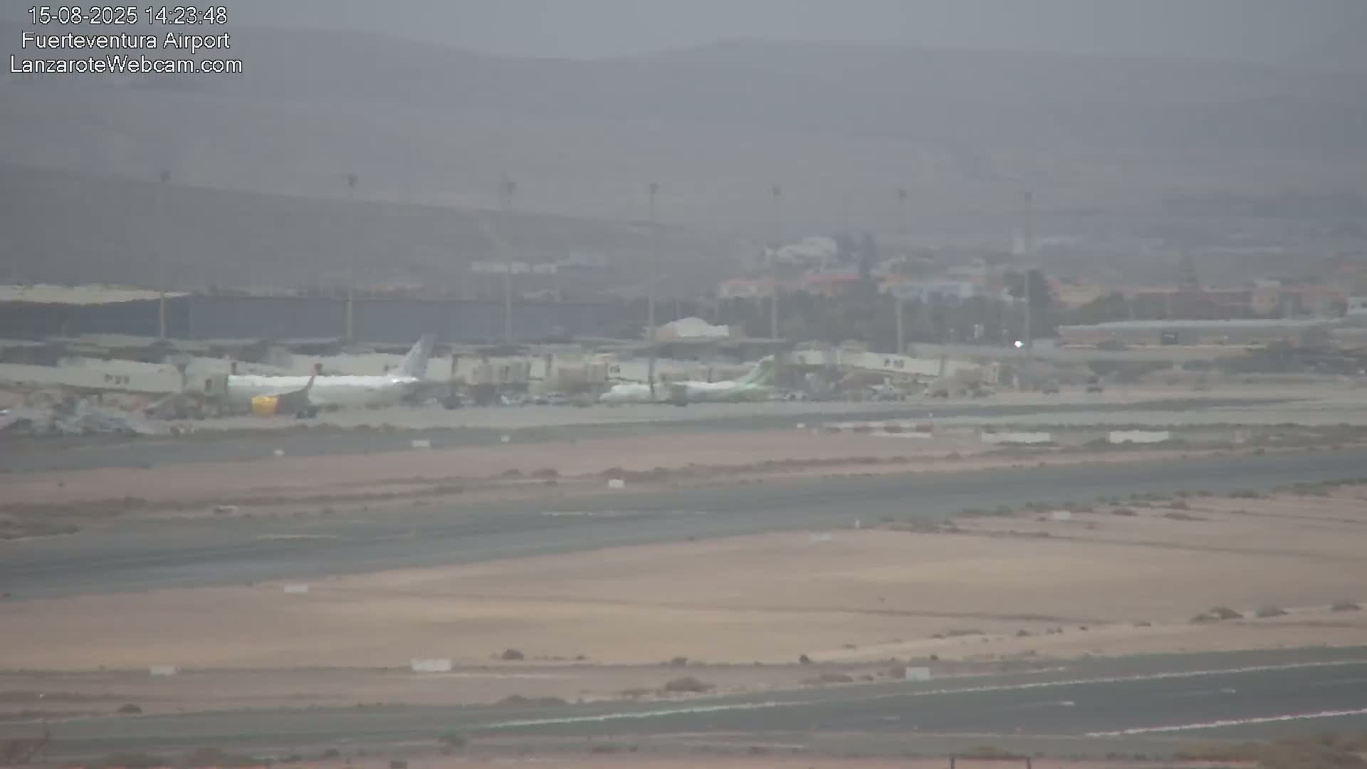 A hazy view of an airport tarmac shows a passenger plane at a gate, with several other gates and airport buildings visible in the background.
