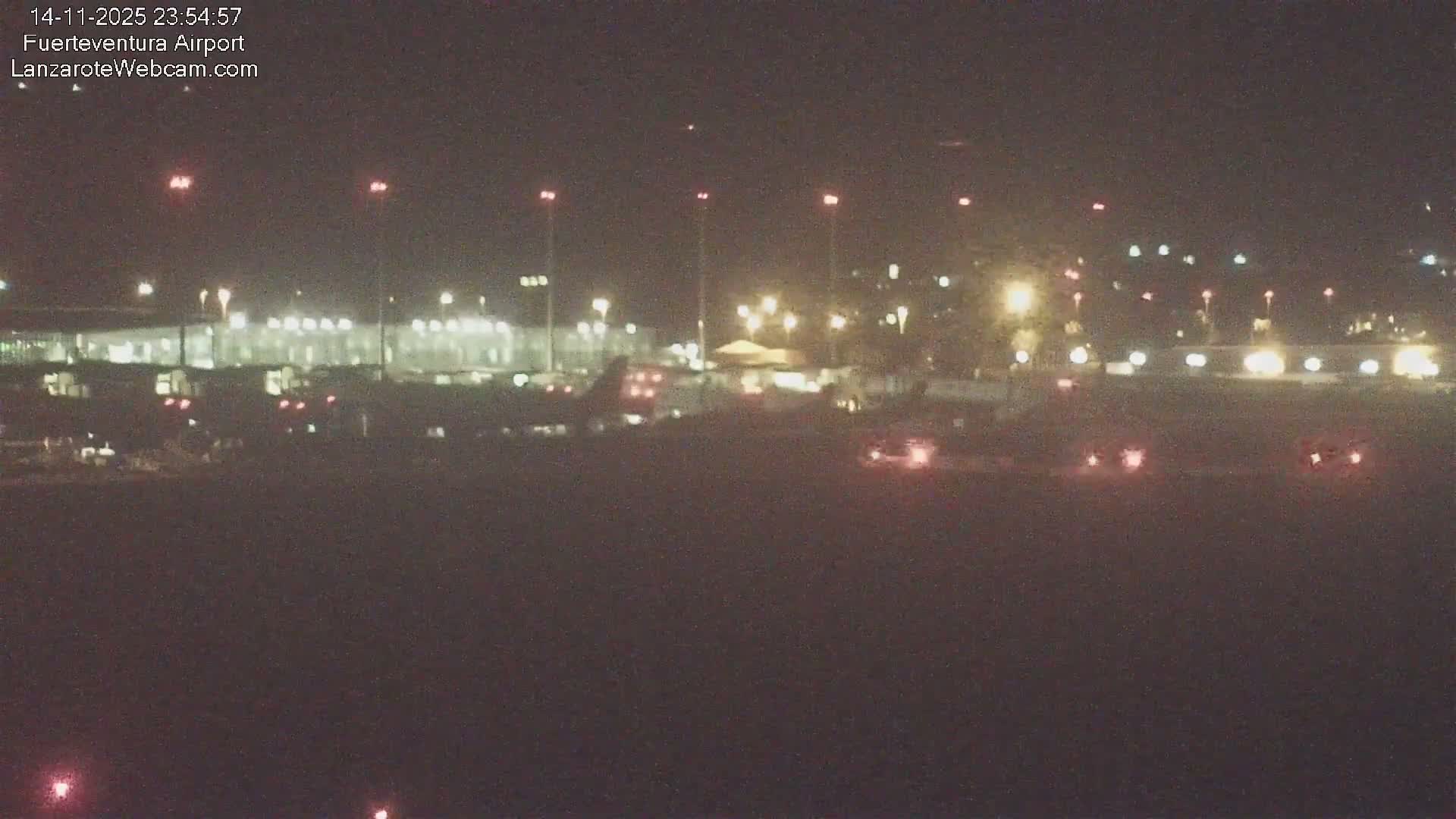 A blurry, low-light view shows an airport at night with a terminal building, parked aircraft, and ground vehicles illuminated by numerous lights under hazy conditions.
