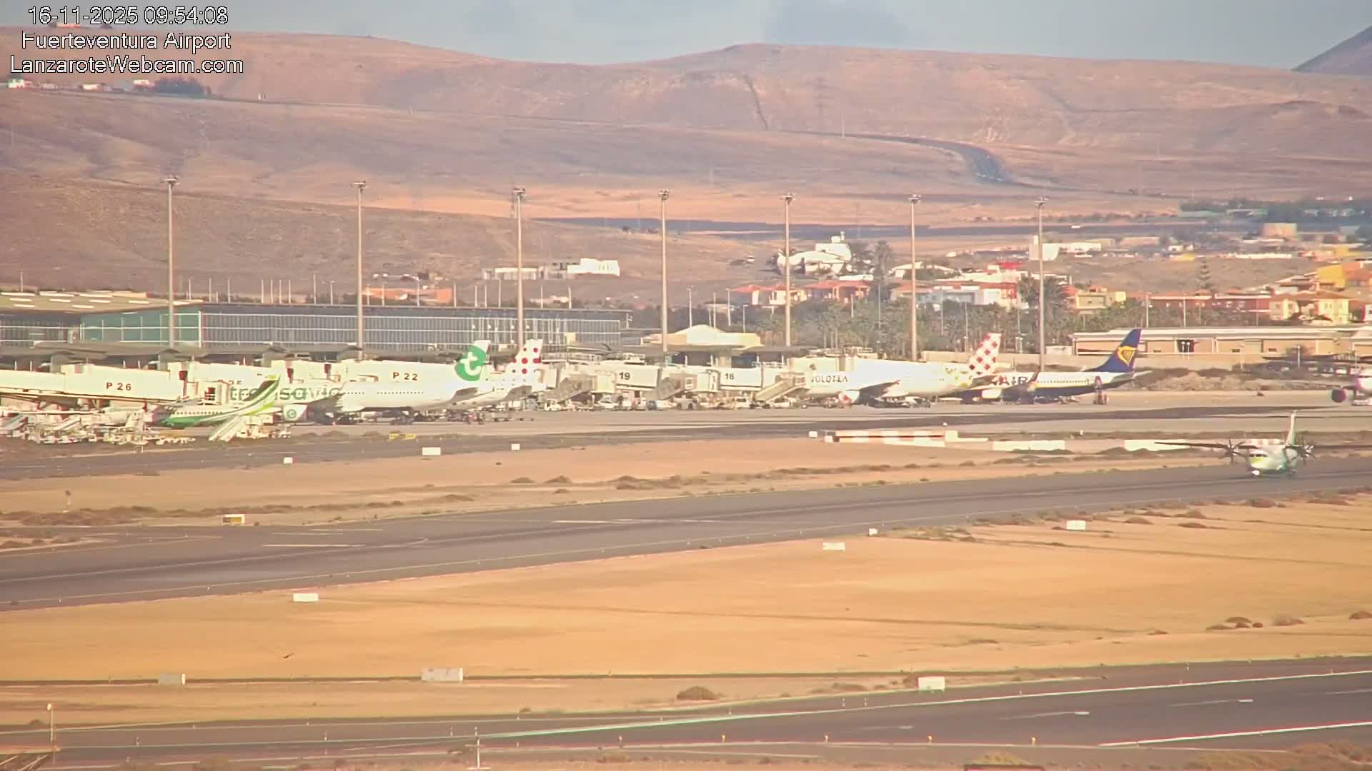 An airport runway and terminal are visible under a clear, sunny sky, with multiple passenger jets at gates and a smaller turboprop plane on a taxiway, all set against a backdrop of arid, distant hills and scattered buildings.