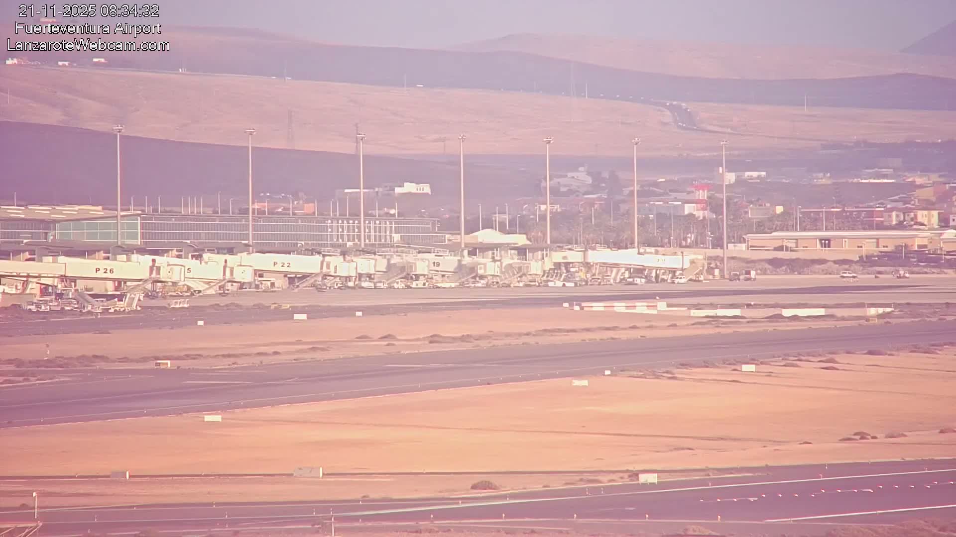 An outdoor view shows an airport tarmac with numerous jet bridges connected to a long terminal building, several runways and taxiways in the foreground, and arid hills rising in the background under clear and fair weather conditions.