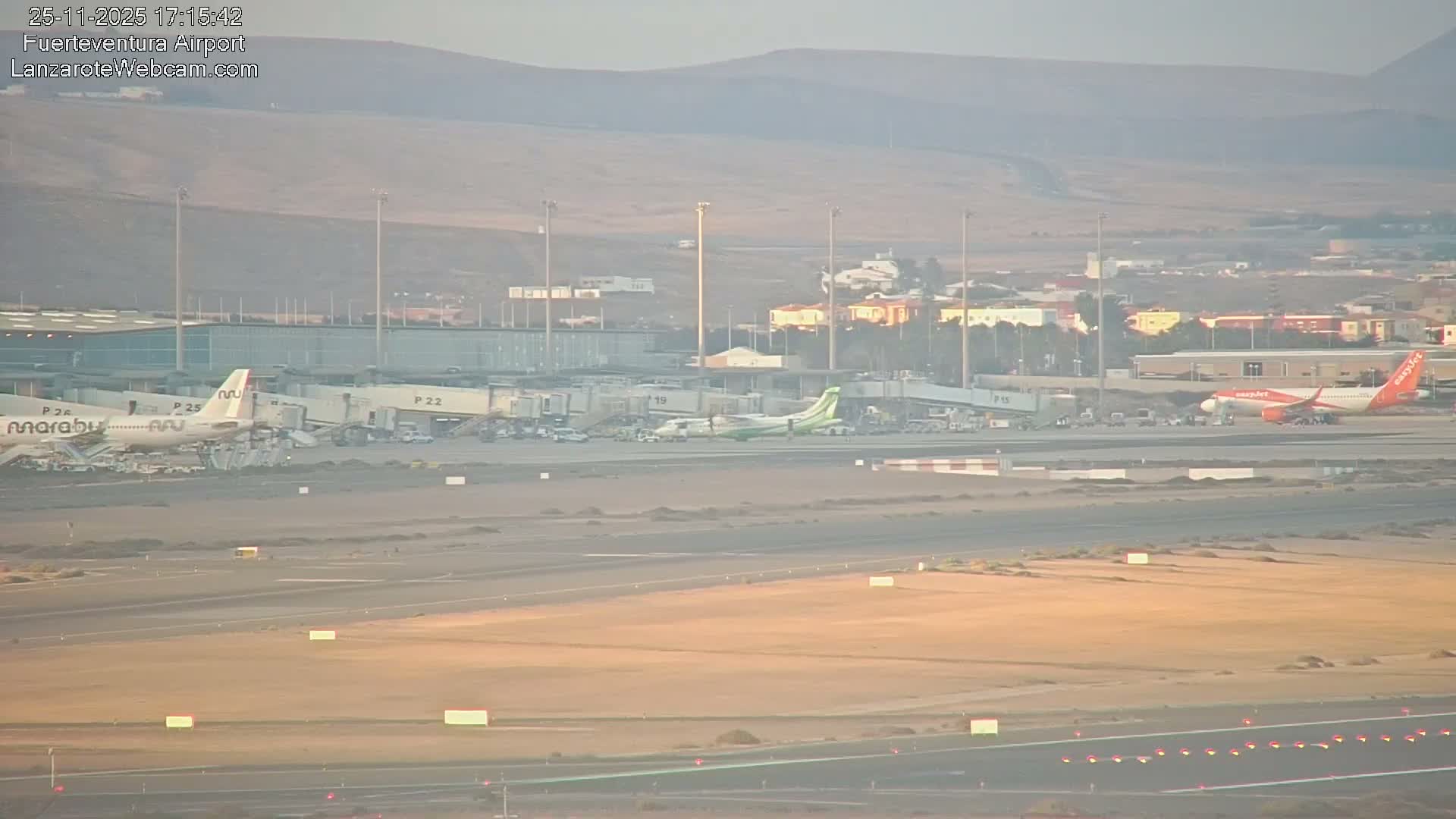 Several airplanes are visible at an airport under hazy, late afternoon skies, with mountains and scattered buildings in the distance beyond the terminal.