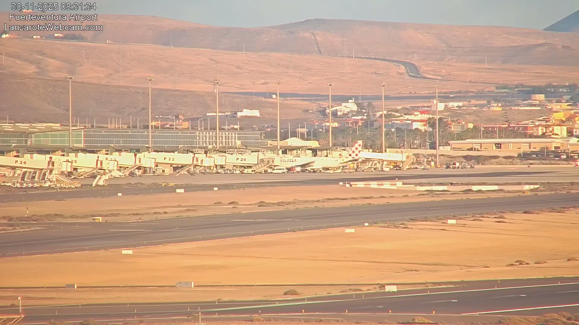 A clear, sunny morning at a desert-like airport features an airplane parked at a gate near a terminal with several jet bridges, all set against a backdrop of arid hills and distant buildings.
