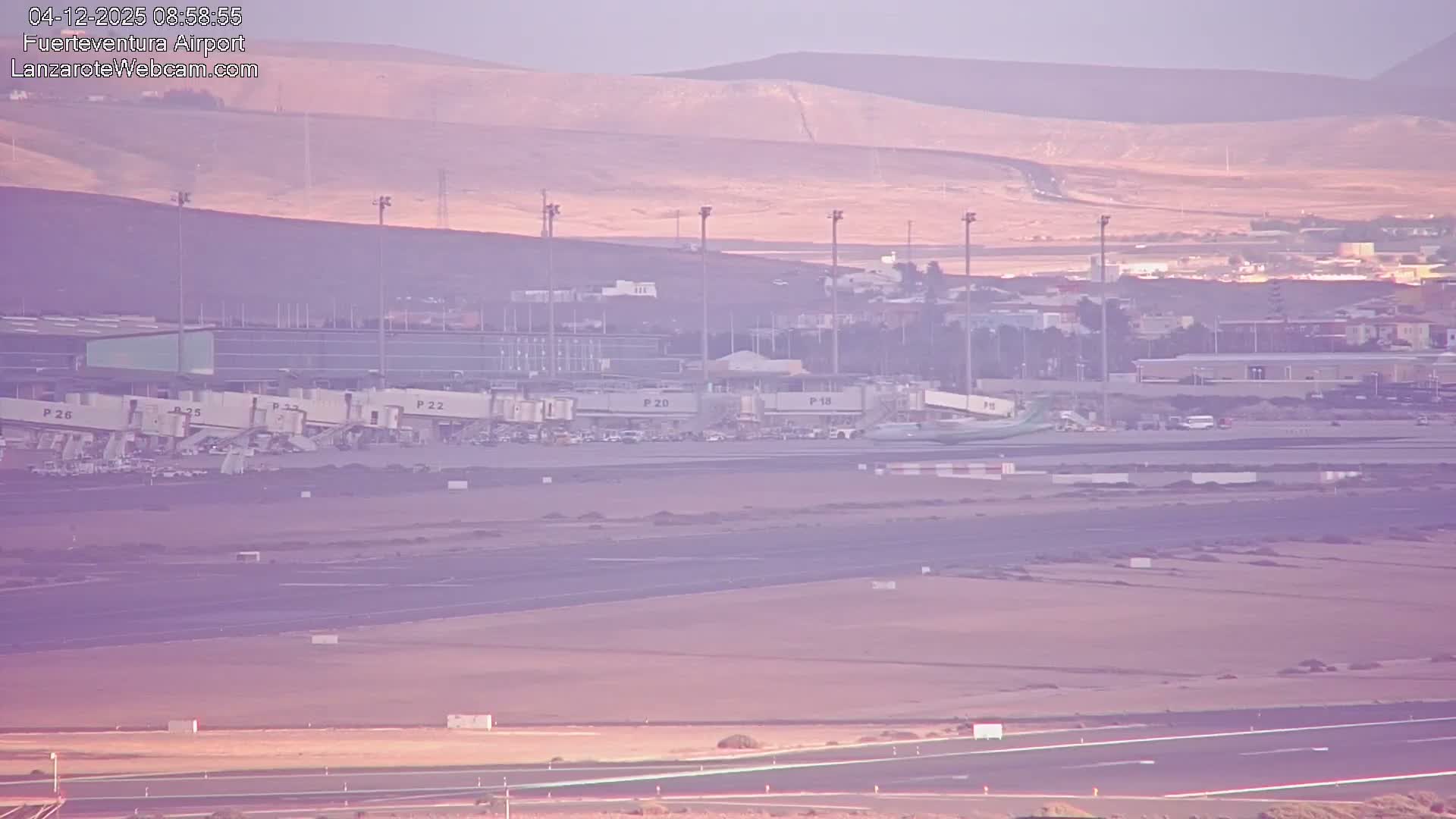 An airport tarmac is visible under a clear, hazy, purple-toned sky, likely at early morning, with several aircraft parked at jet bridges, various ground vehicles, and runways stretching into the foreground, all set against a backdrop of arid, undulating hills.