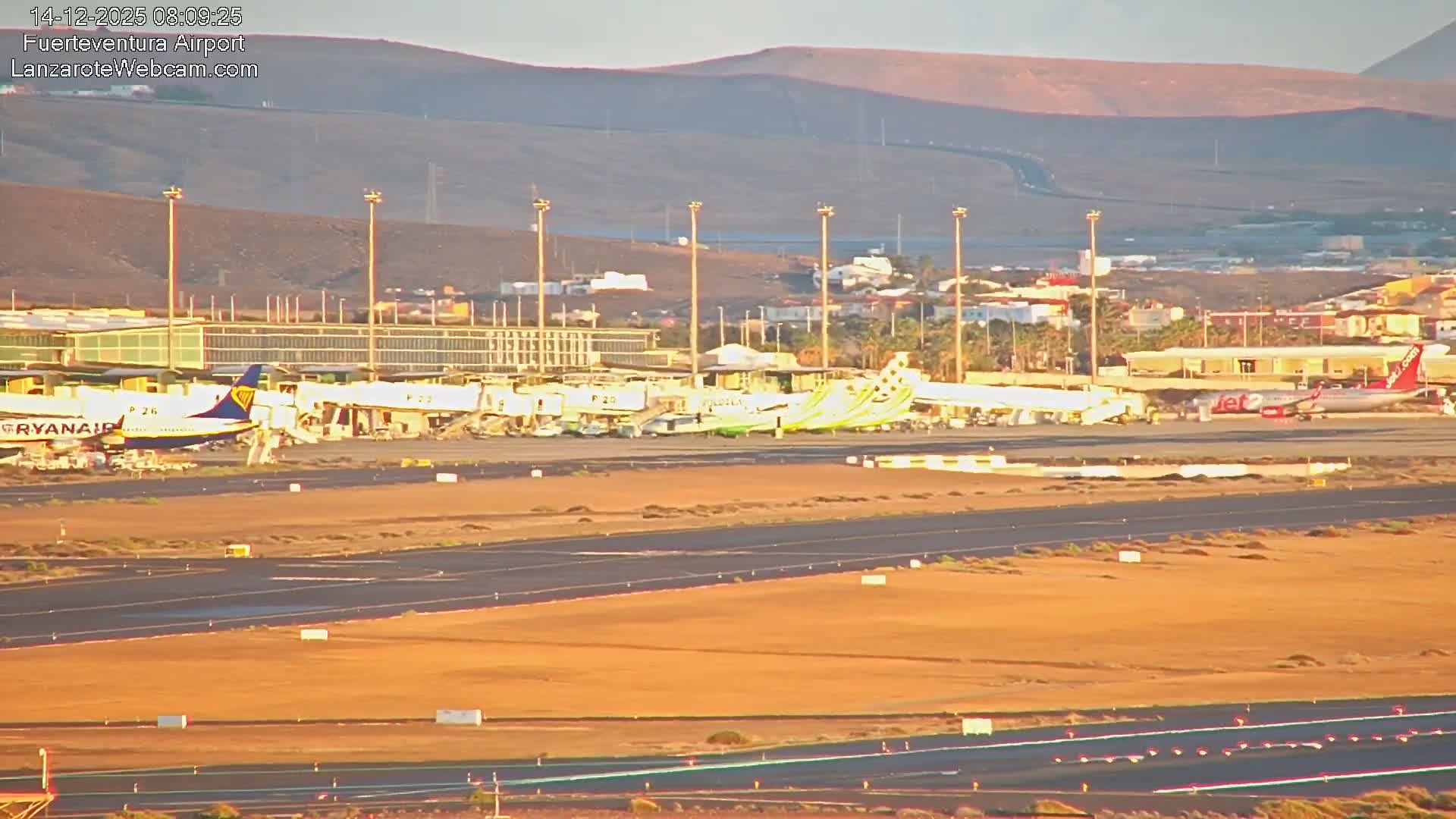 The image shows a bustling airport on a clear, sunny day, with several passenger airplanes parked at jet bridges alongside terminal buildings, and runways extending into a dry, hilly landscape under a bright sky.