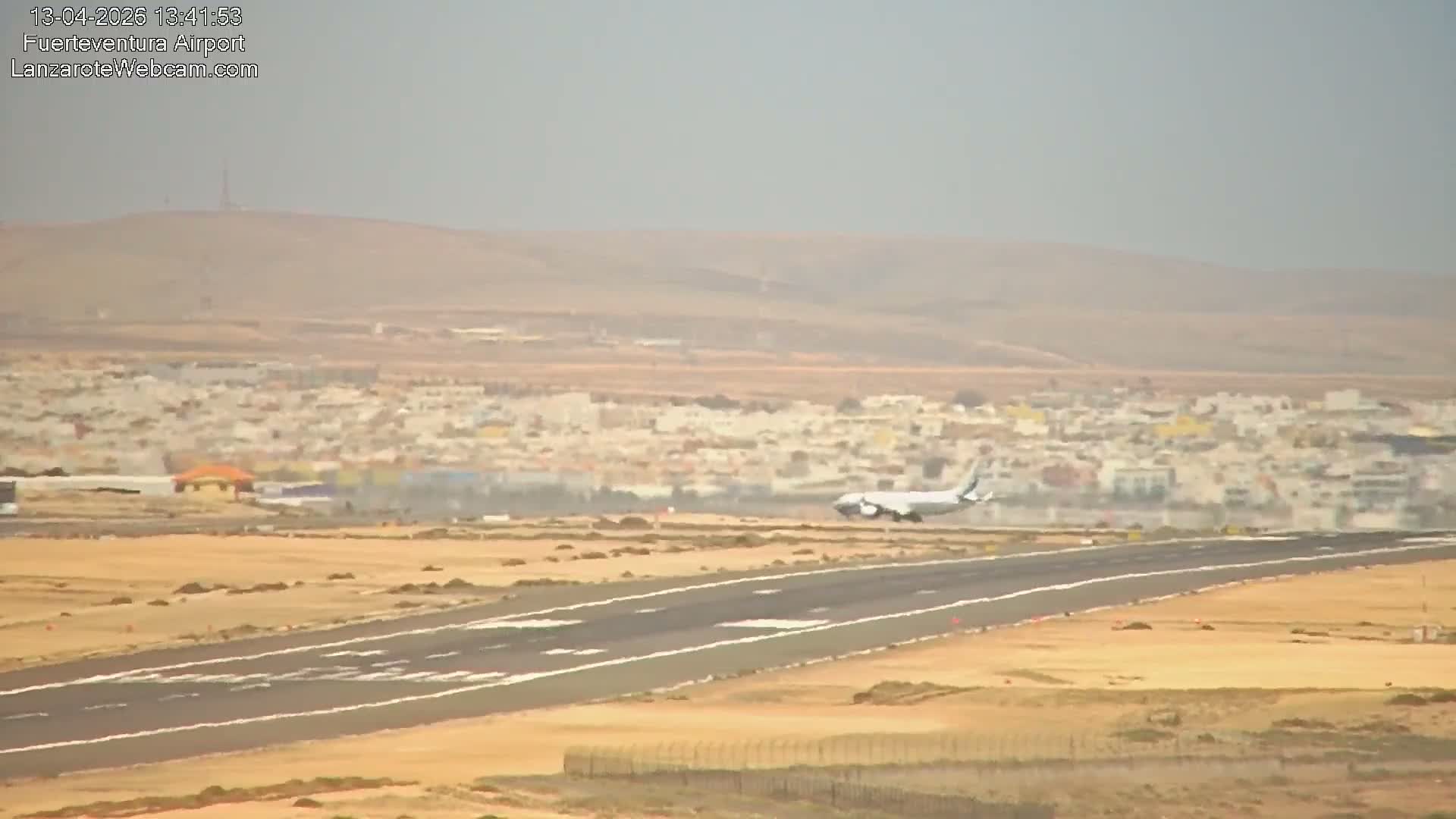 The image shows a bustling airport on a clear, sunny day, with several passenger airplanes parked at jet bridges alongside terminal buildings, and runways extending into a dry, hilly landscape under a bright sky.