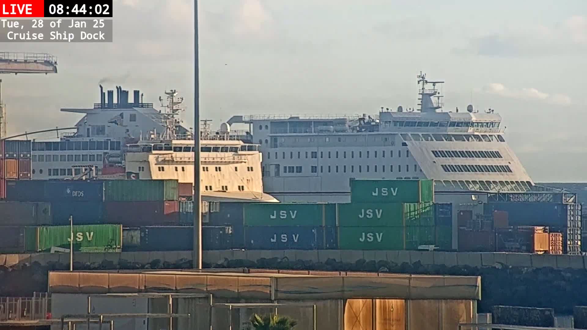 A large, white ferry is docked at a port, surrounded by stacked shipping containers under a mostly clear sky.