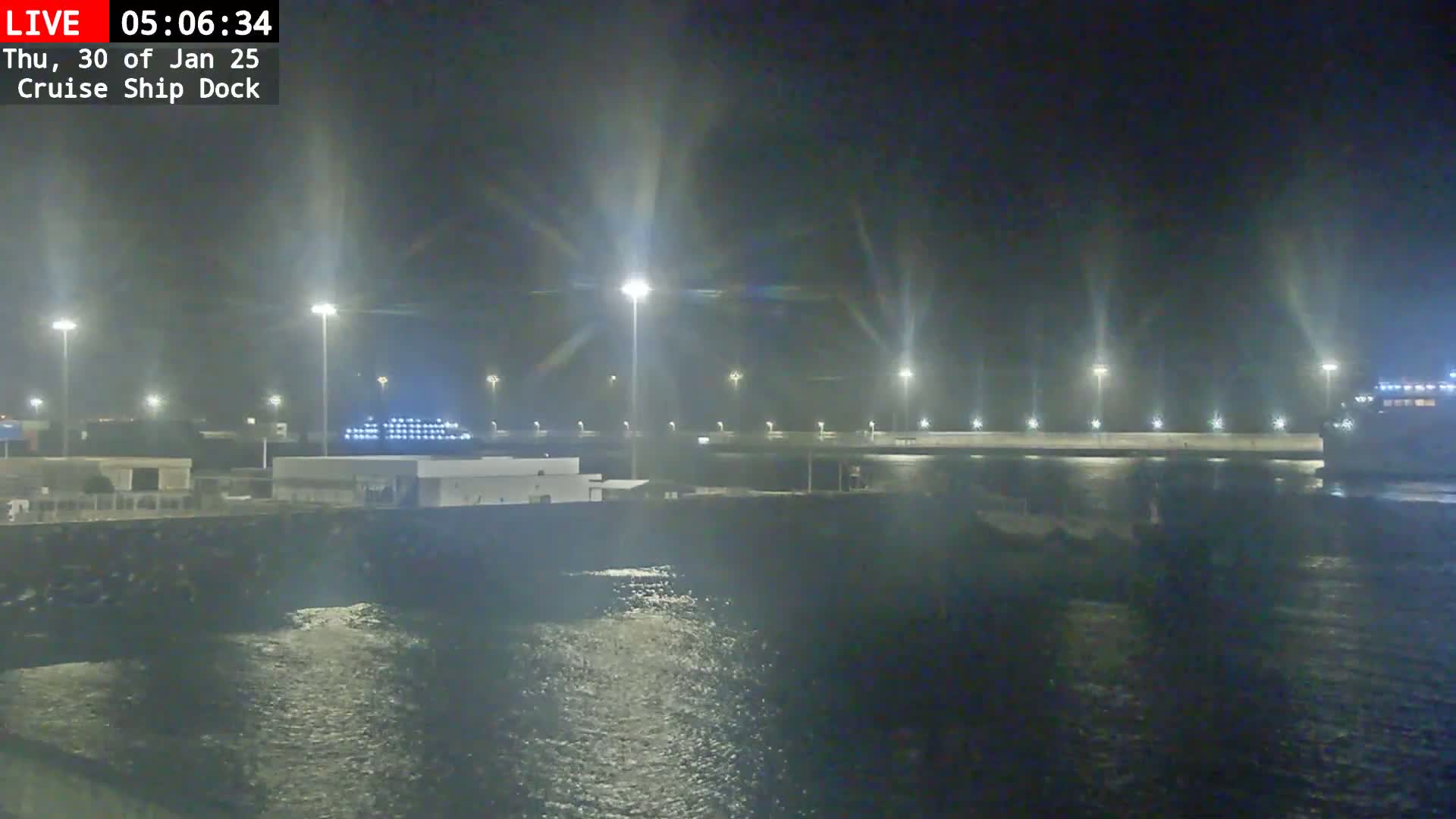 A nighttime view of a cruise ship docked at a port, with calm water and bright lights illuminating the scene.