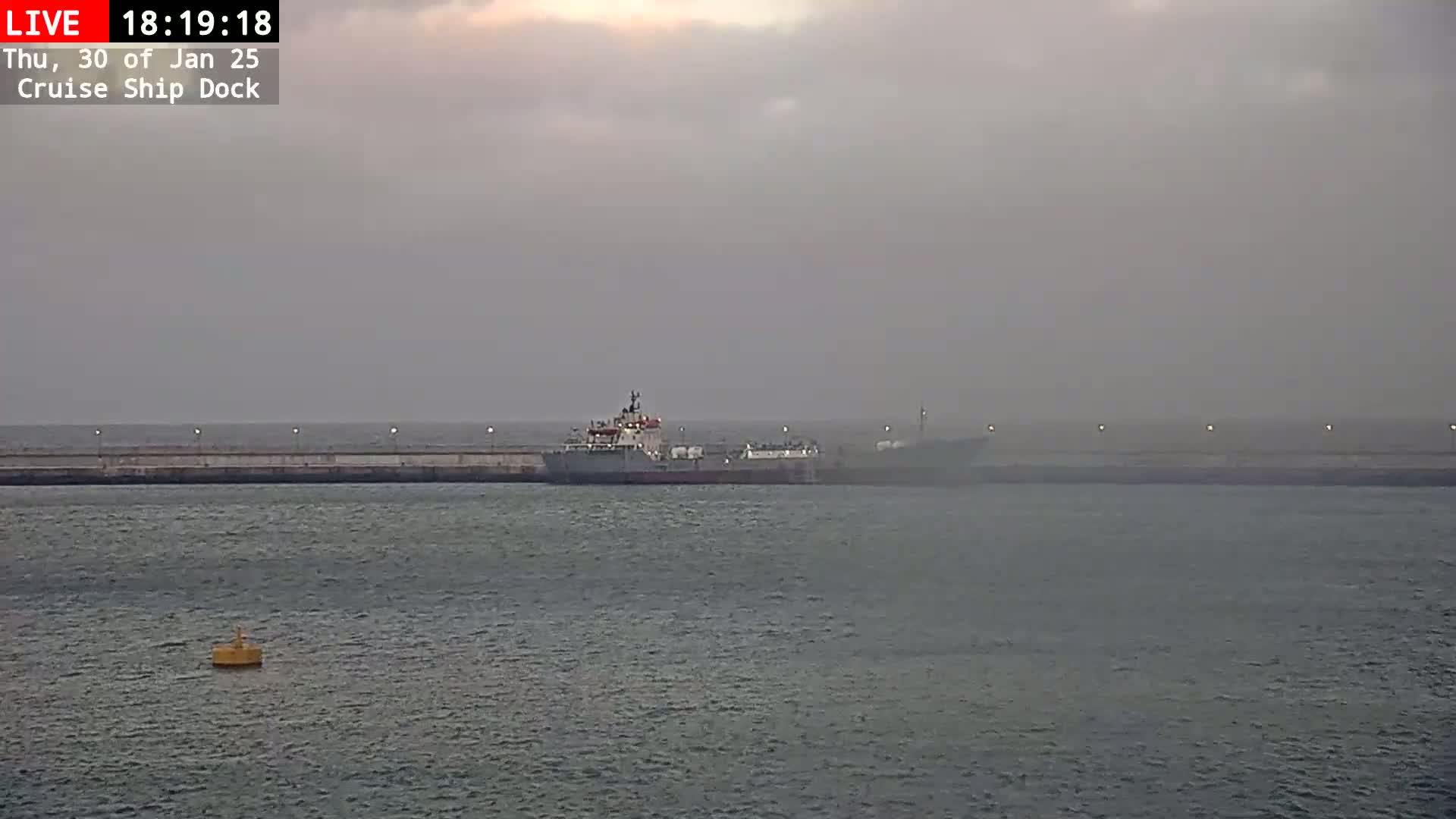 A hazy, overcast day shows a large ship docked at a pier with a smaller vessel alongside it.