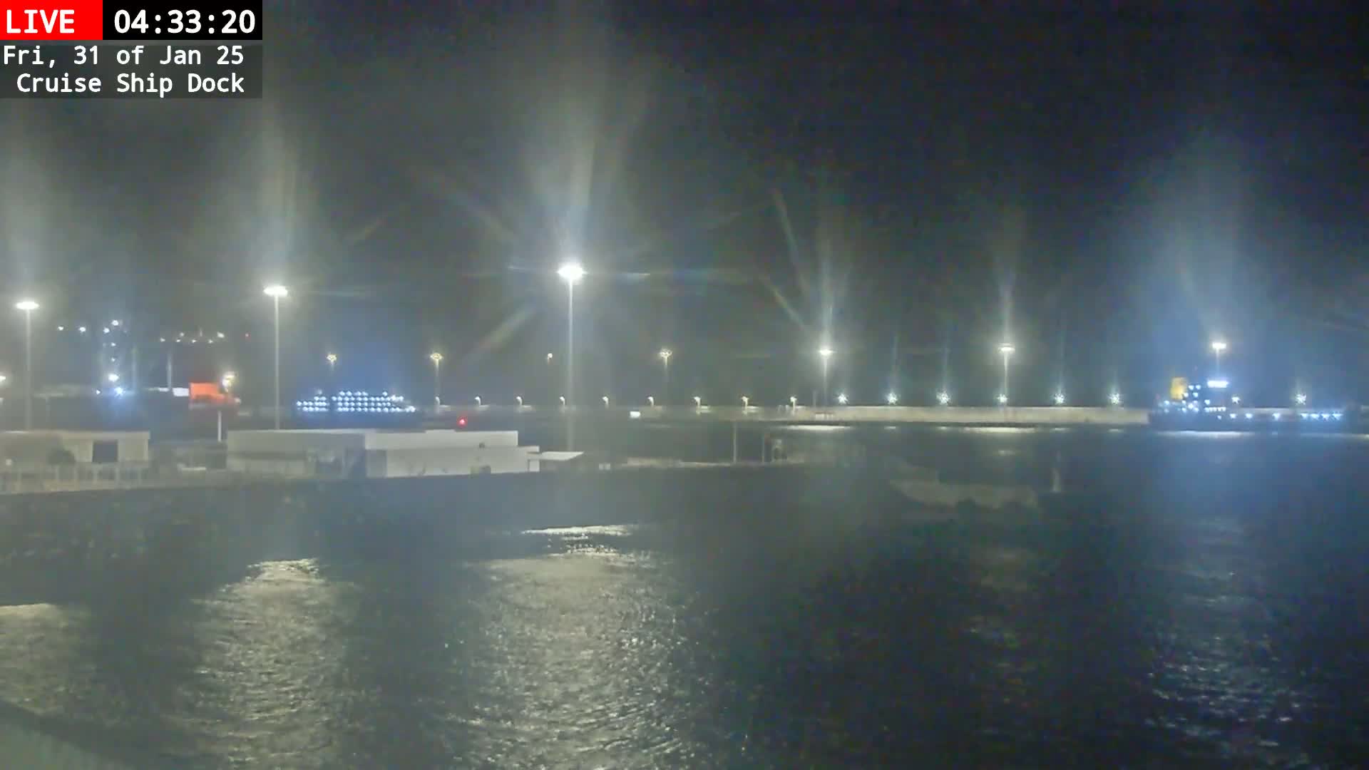 A nighttime view of a cruise ship docked at a port, with calm water and bright lights illuminating the scene.