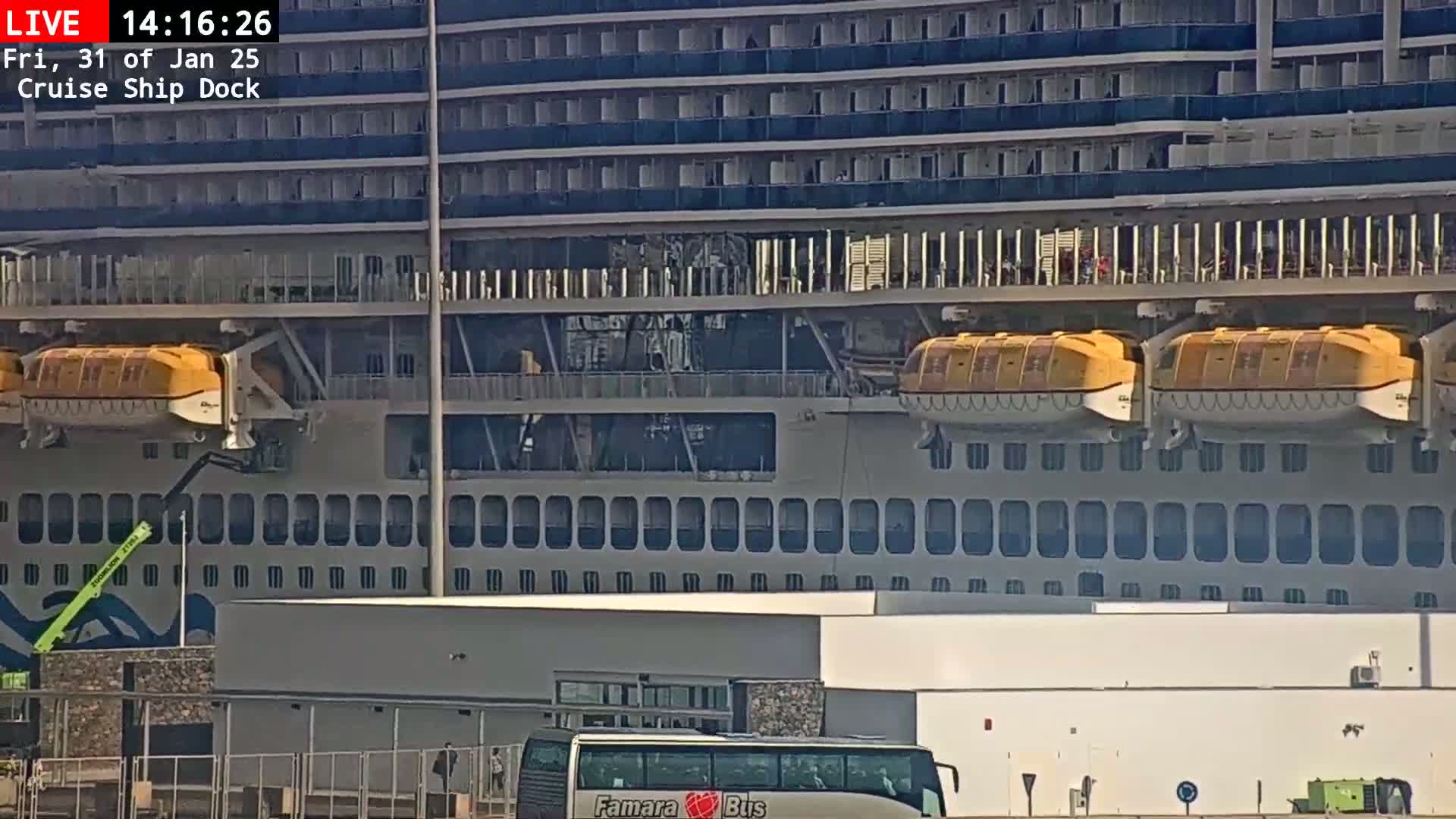 A large cruise ship is docked, with two yellow lifeboats visible, under sunny conditions.