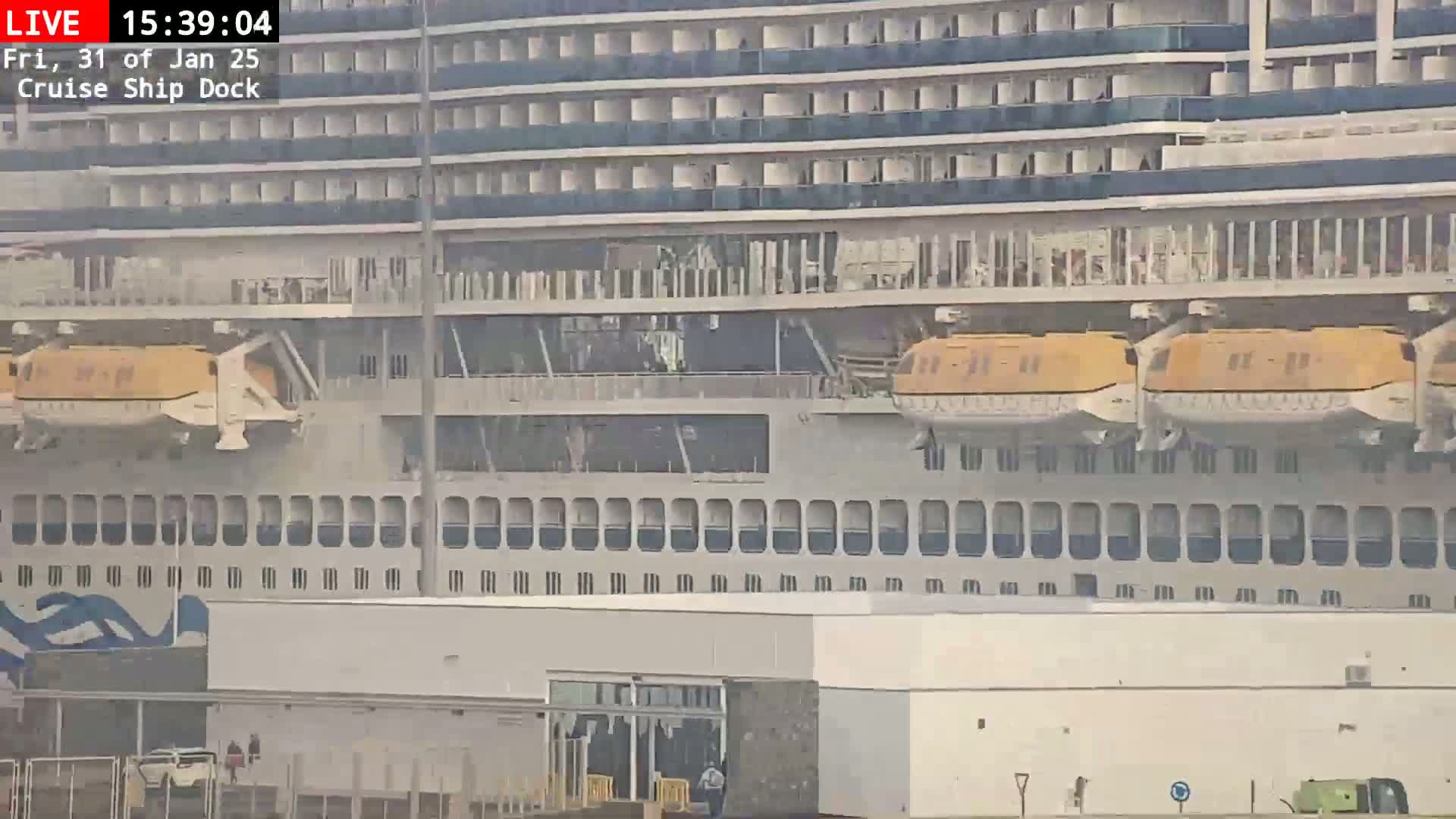 A large cruise ship is docked, with two orange lifeboats visible on its side, in overcast conditions.