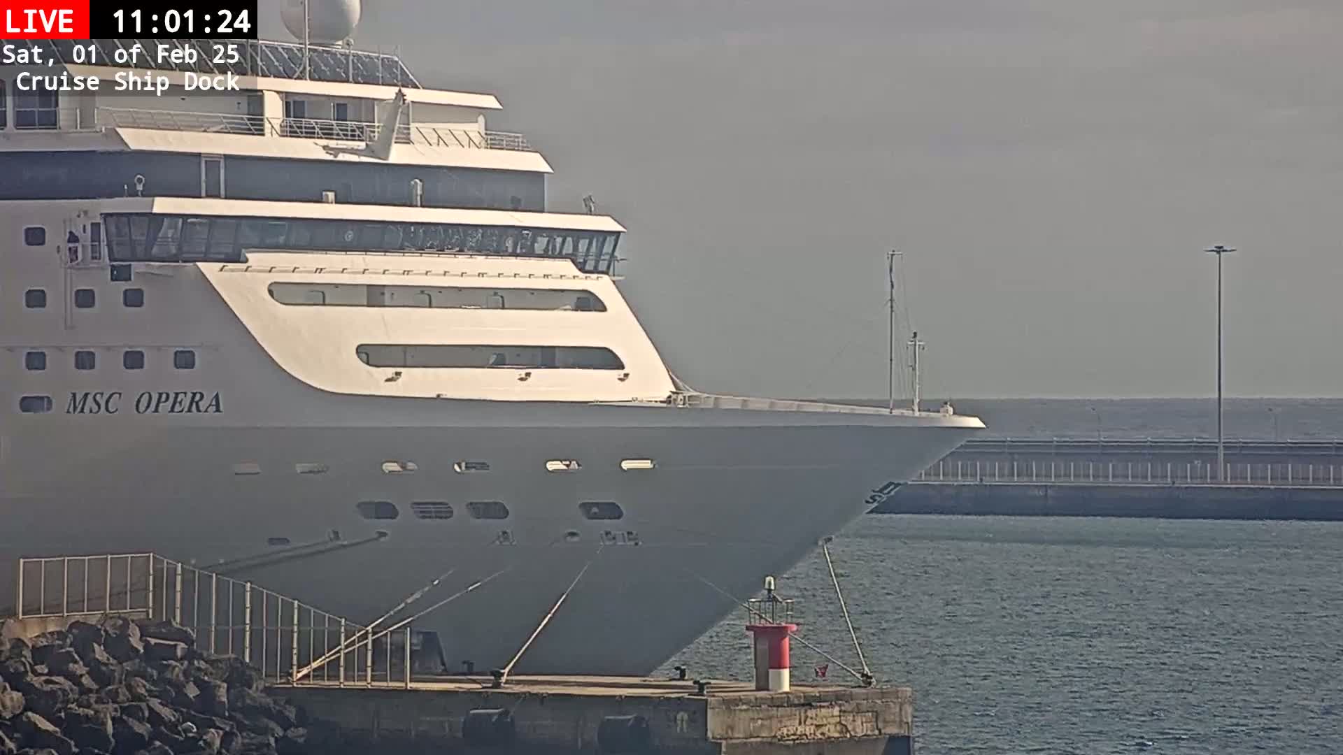 The bow of a large cruise ship, named MSC Opera, is docked at a pier on a calm, hazy day.