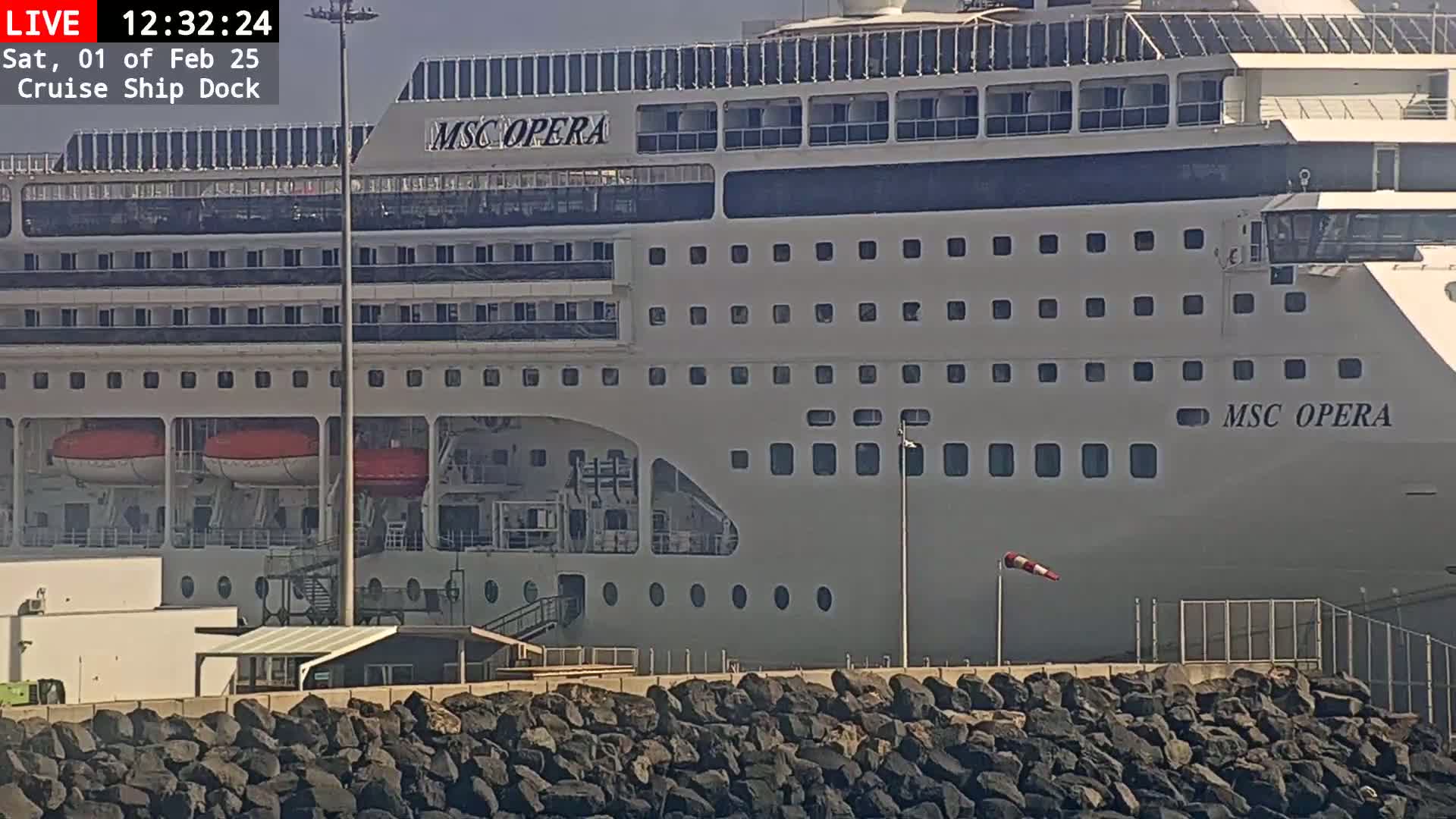 A large cruise ship is docked at a port on a sunny day, with a rock wall separating the ship from the land.
