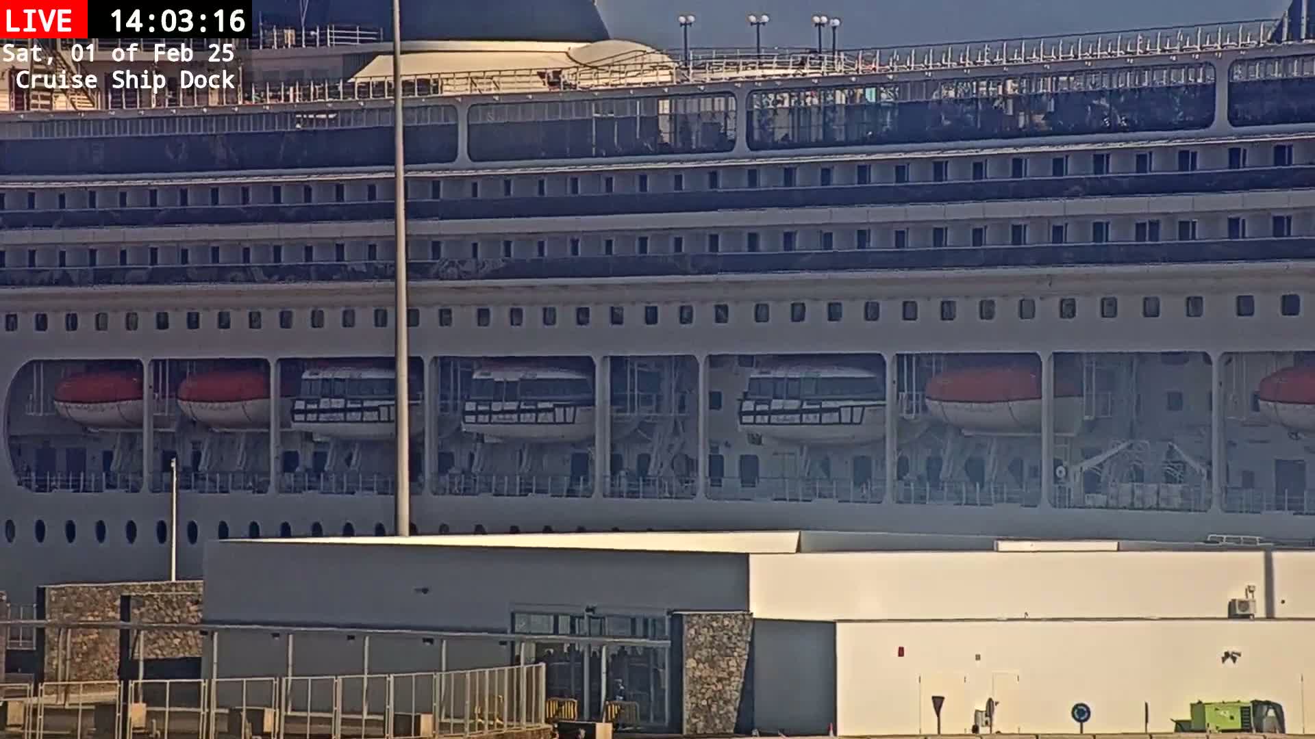 A large cruise ship is docked at a port on a sunny day, with several lifeboats visible on its side.