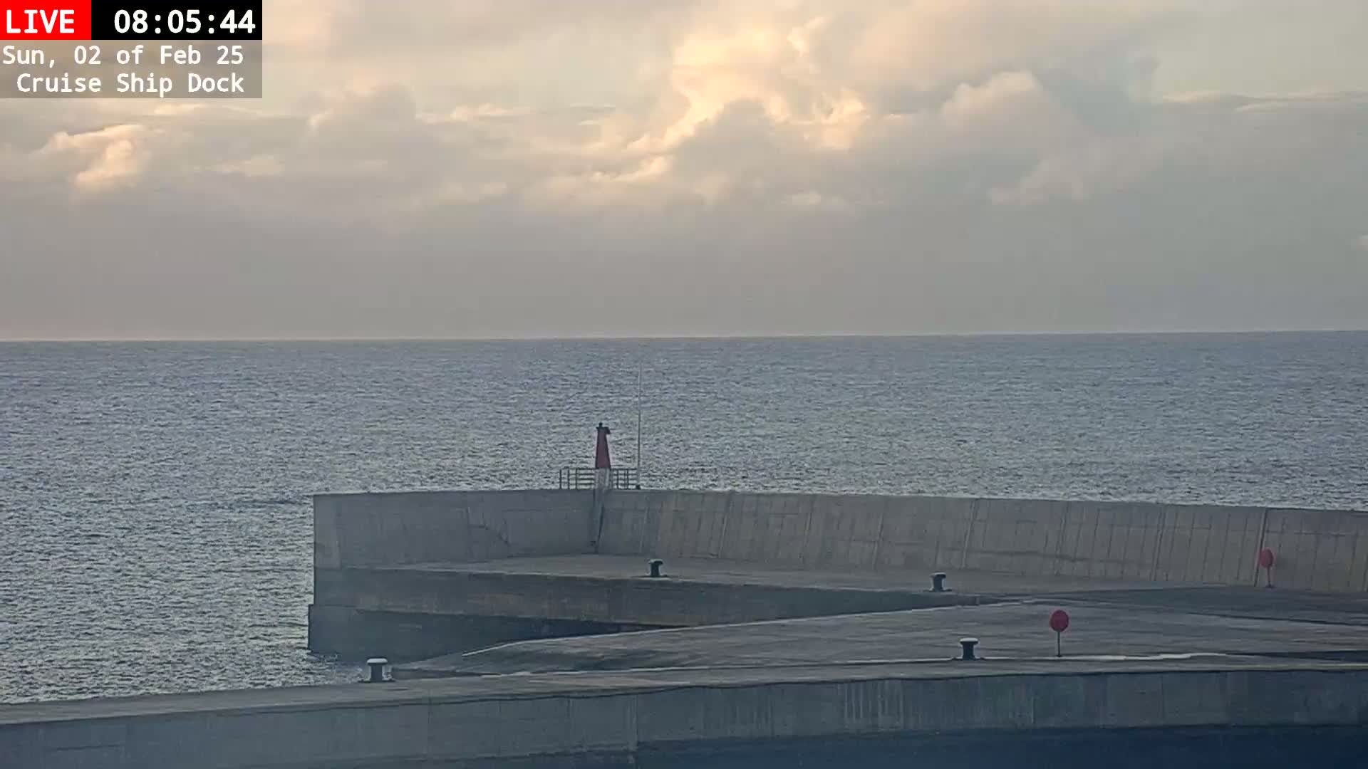 A calm sea is visible under a partly cloudy sky, with a concrete cruise ship dock extending into the water, featuring a small lighthouse.
