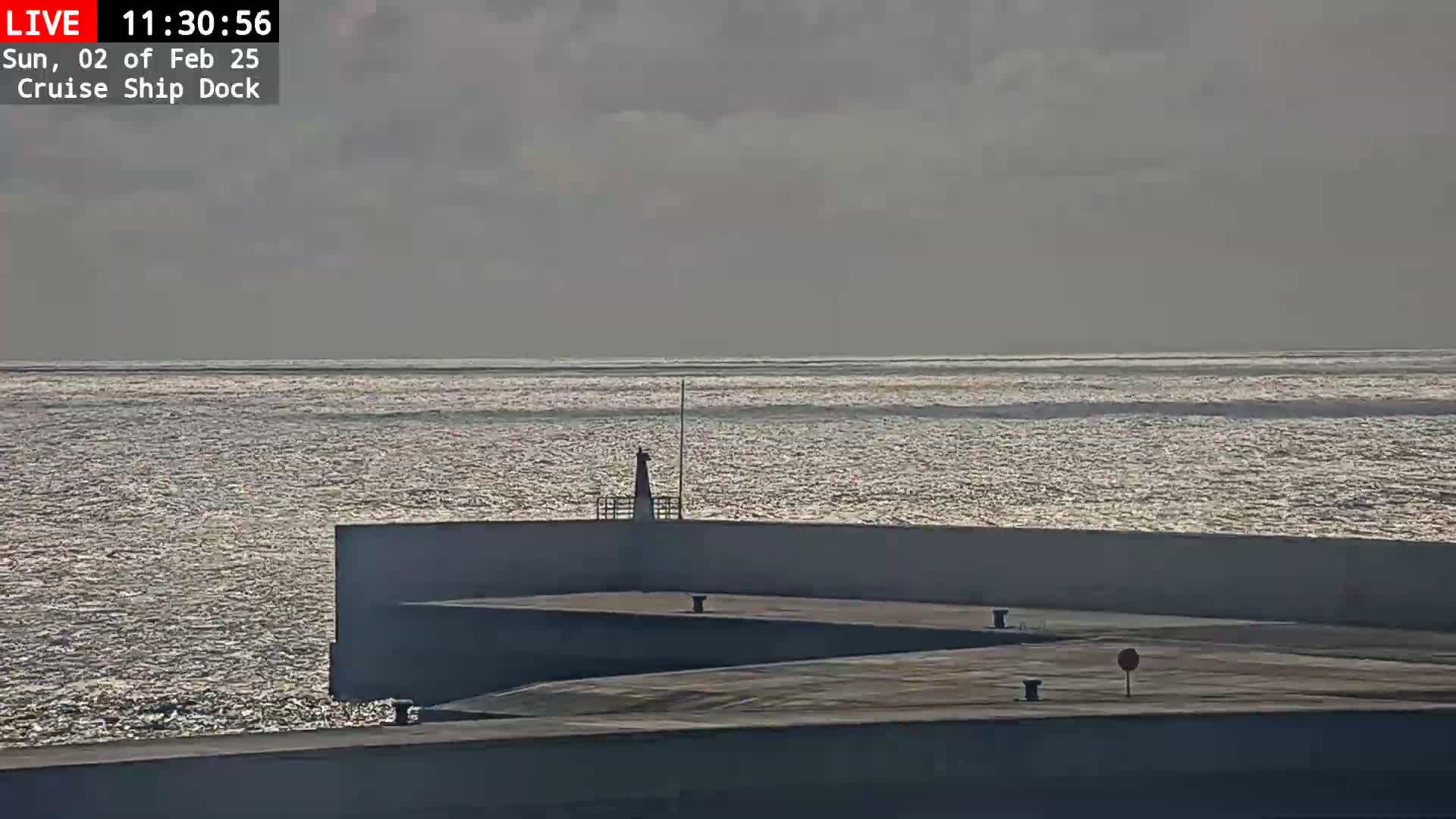 A calm, glittering ocean stretches to the horizon under an overcast sky, viewed from a cruise ship dock with a low wall and small structures.