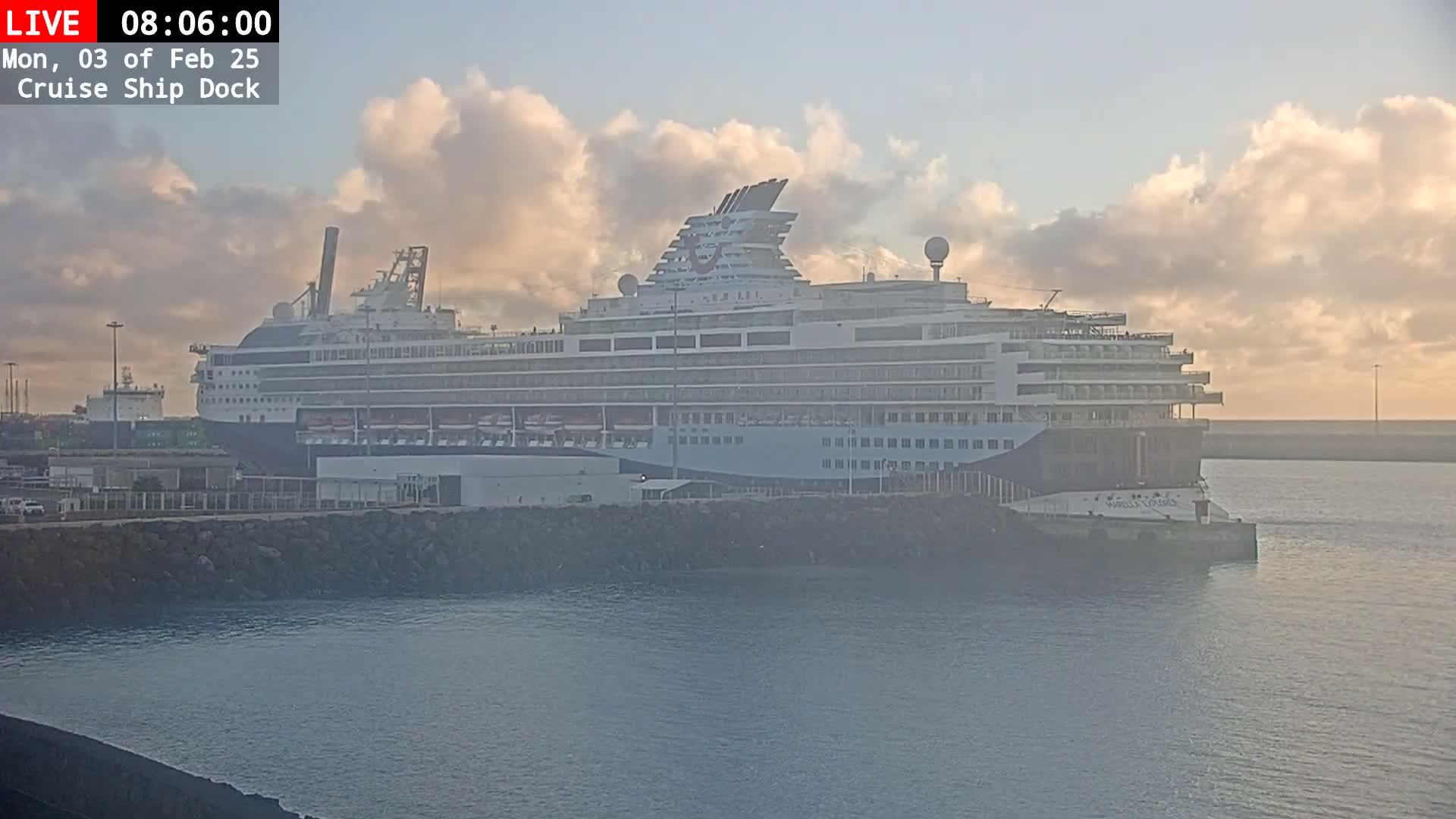 A large cruise ship is docked at a pier under a partly cloudy sunrise.