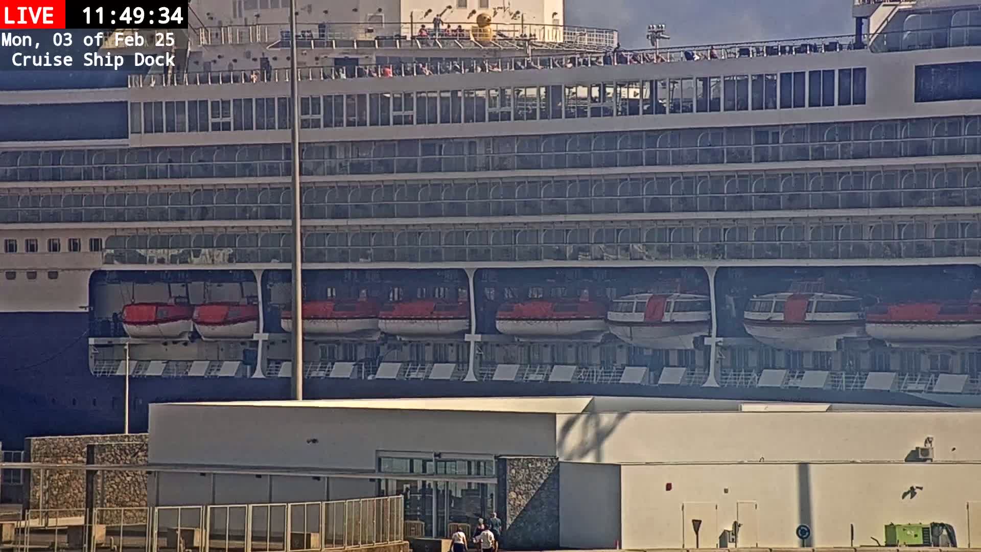 A large cruise ship is docked at a port on a sunny day, with several lifeboats visible on its hull.