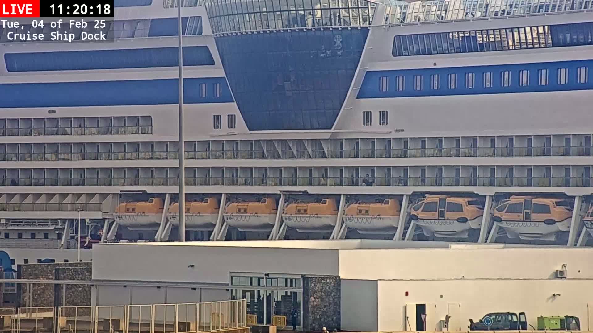 A large cruise ship with multiple orange lifeboats is docked at a port on a partly cloudy day.