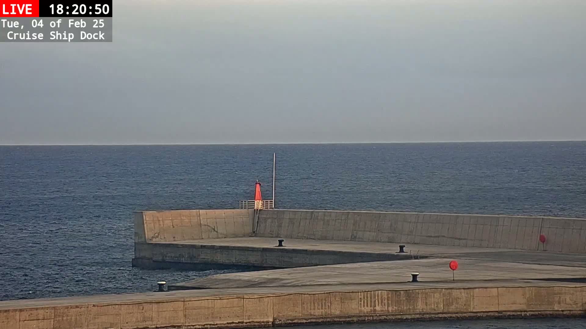 A concrete cruise ship dock extends into a calm, dark blue sea under a hazy sky, with a small red lighthouse visible on the dock.