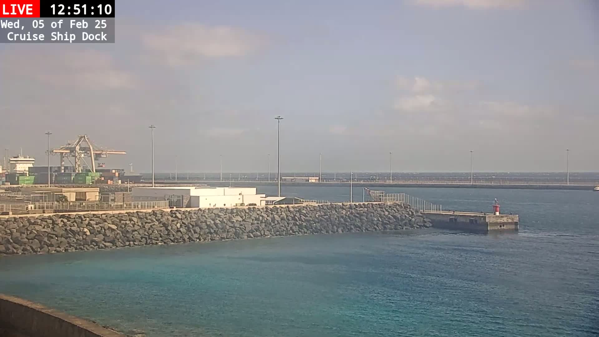 A cruise ship dock on a partly cloudy day shows a cargo ship being unloaded by a crane, a rocky seawall, and calm blue water.