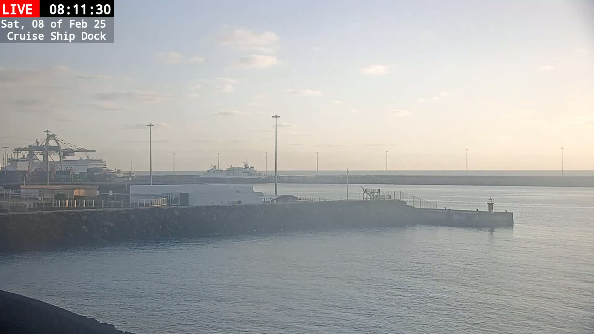 A hazy sunrise illuminates a cruise ship dock with several ships, including a large cruise ship and cargo ships, docked alongside a protective wall extending into calm water.