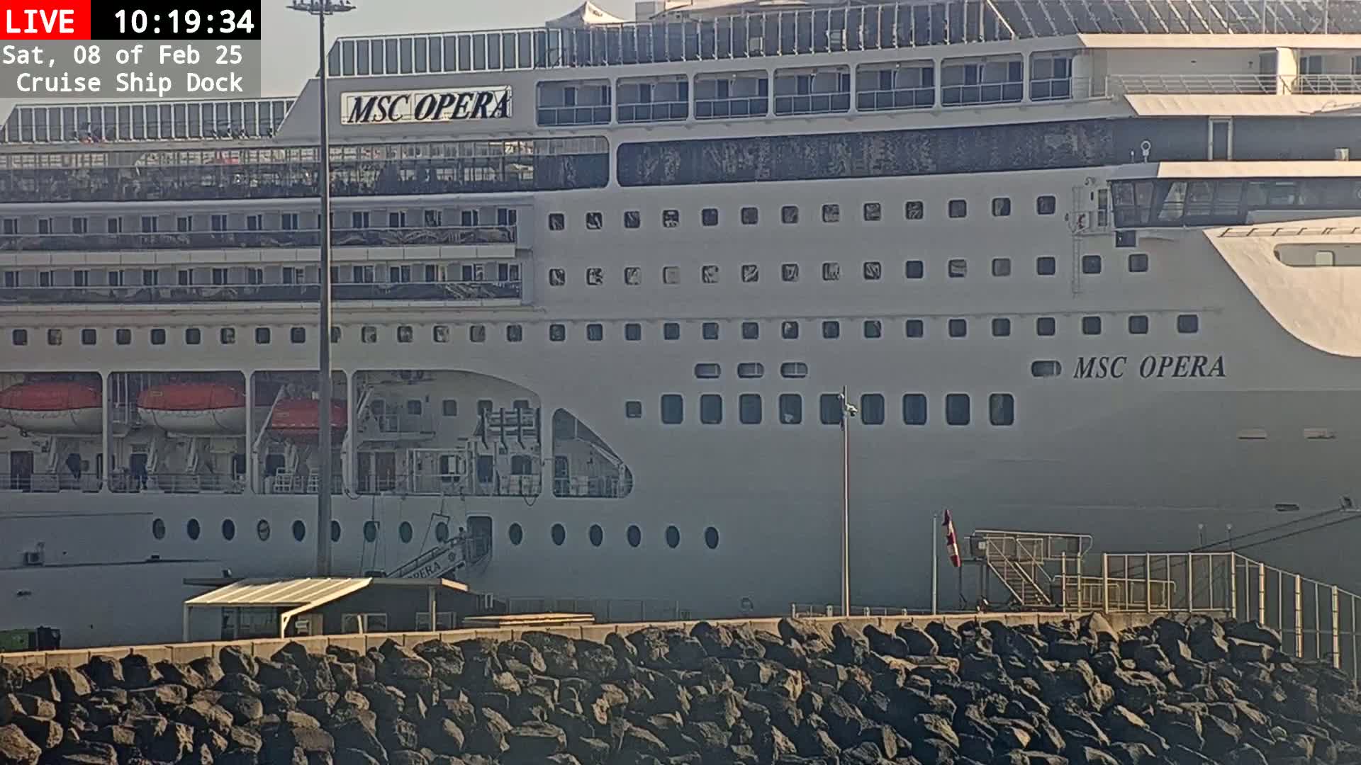A large cruise ship, the MSC Opera, is docked at a pier on a sunny day, with a rocky embankment in the foreground.