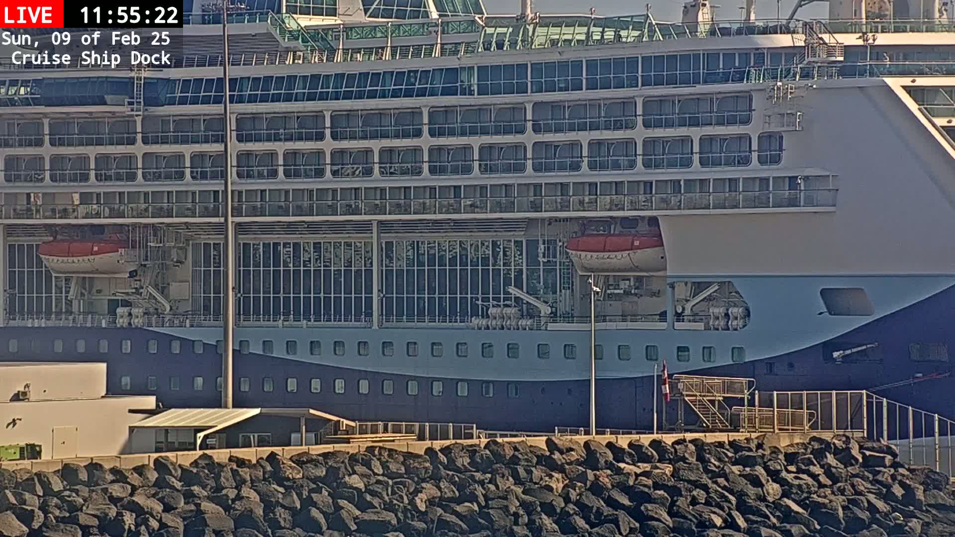 A large cruise ship is docked at a pier on a sunny day, with a rocky embankment in the foreground.