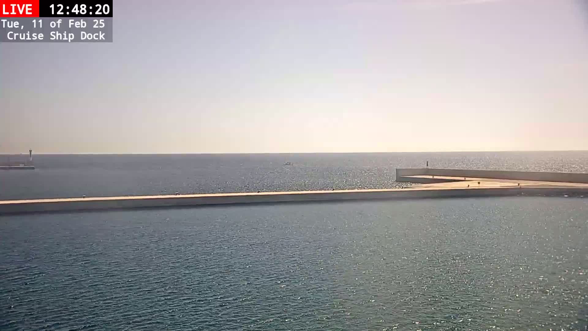 A long, light-colored pier extends into a calm, blue sea under a hazy sky.