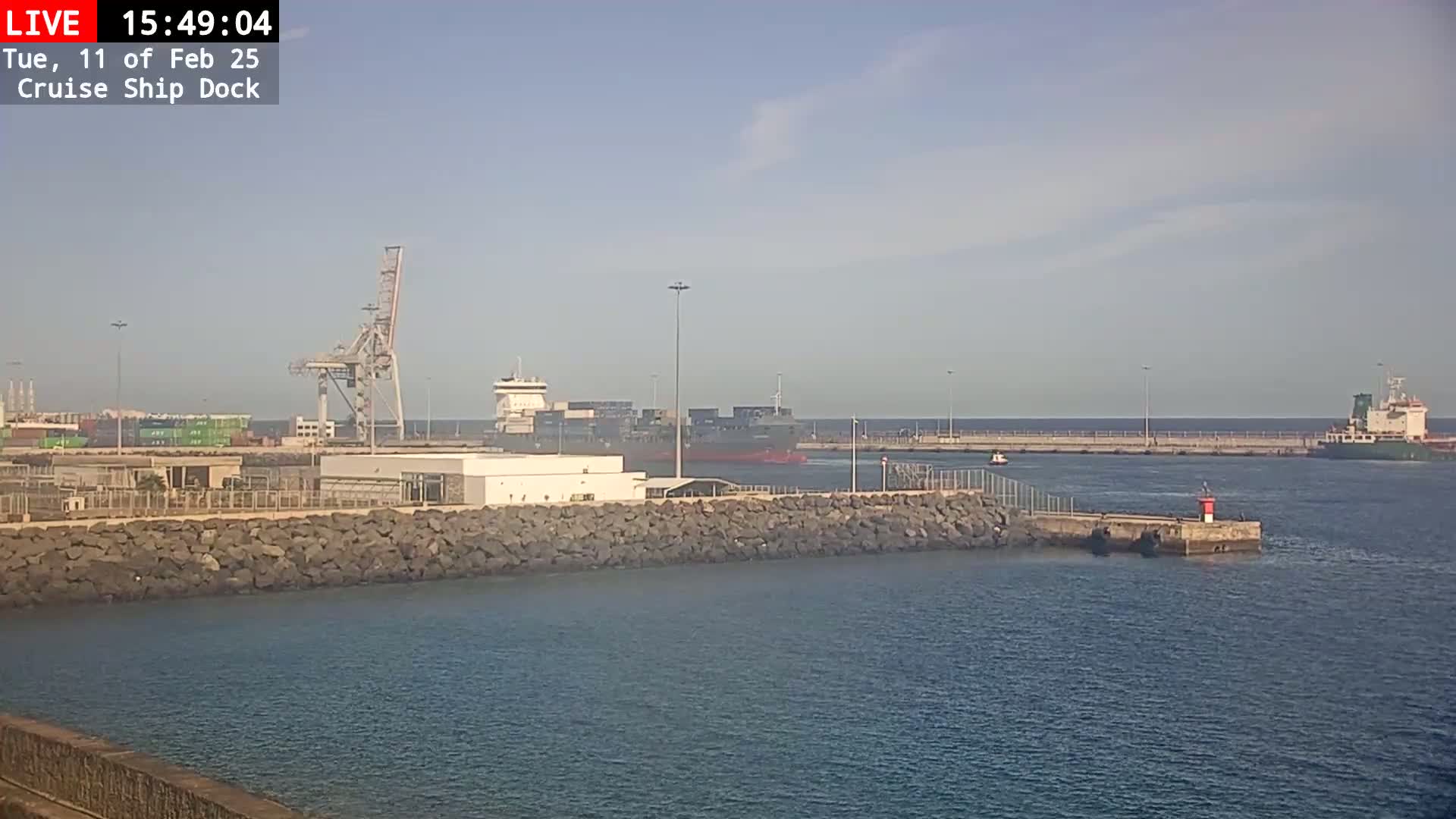 A container ship is docked at a port on a calm, sunny day, with another ship visible further out in the water.