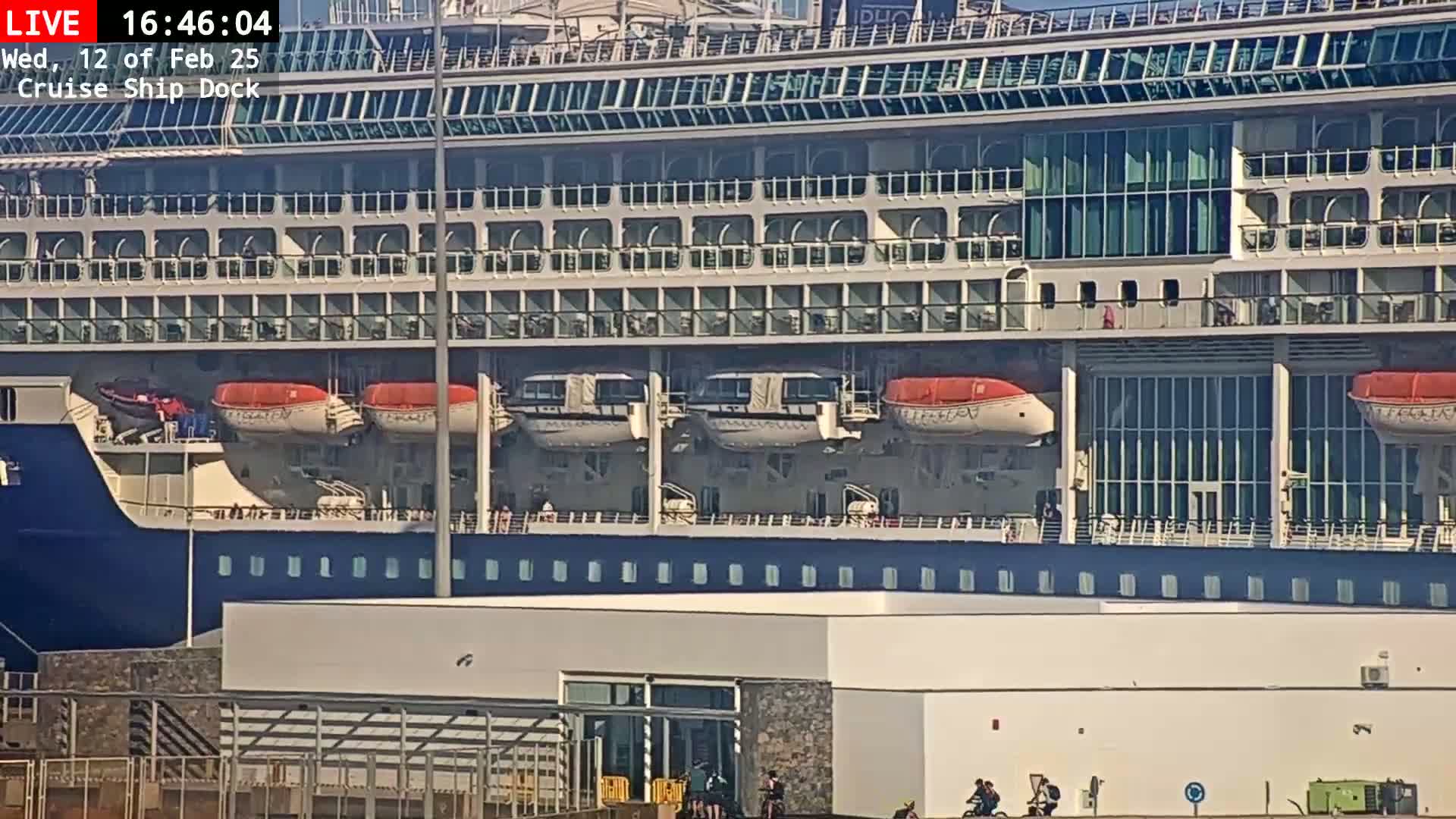 A large cruise ship is docked at a port on a sunny day, with several lifeboats visible on its side.