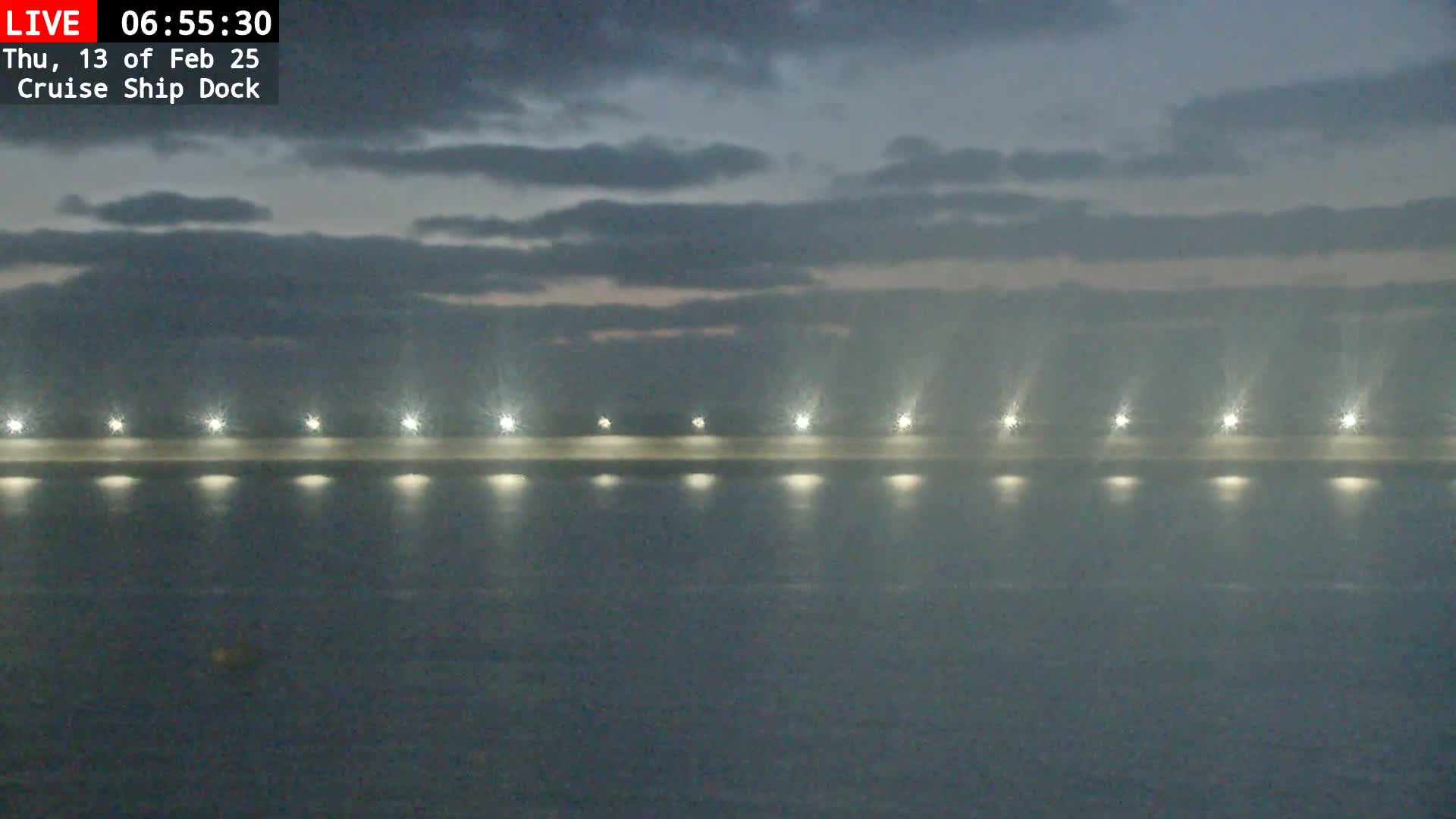 A cruise ship dock at night under a cloudy sky is lit by several lights along its length, reflecting on the dark water.