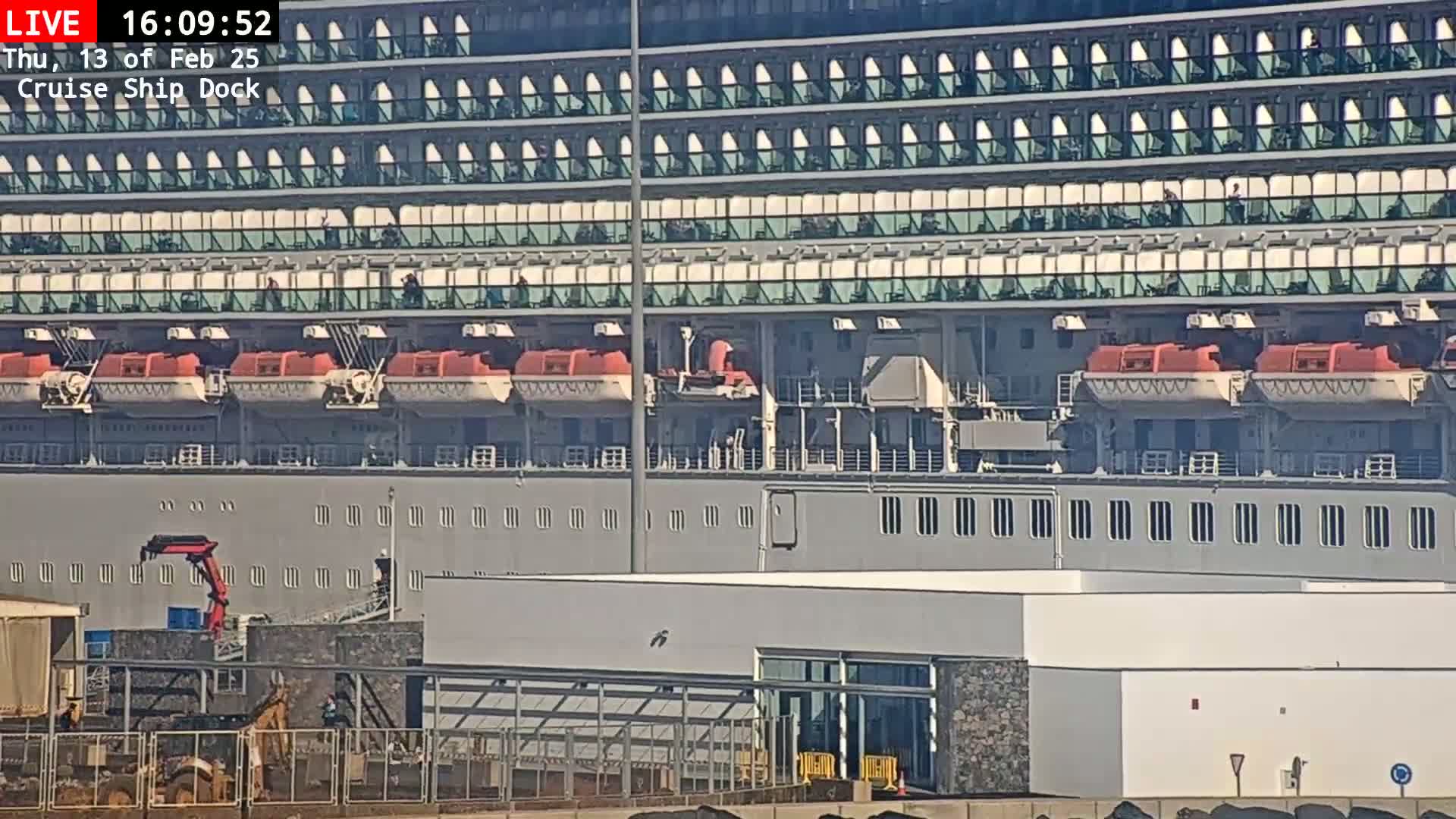 A large cruise ship is docked at a port on a sunny day, with a crane and other equipment visible on the dock.
