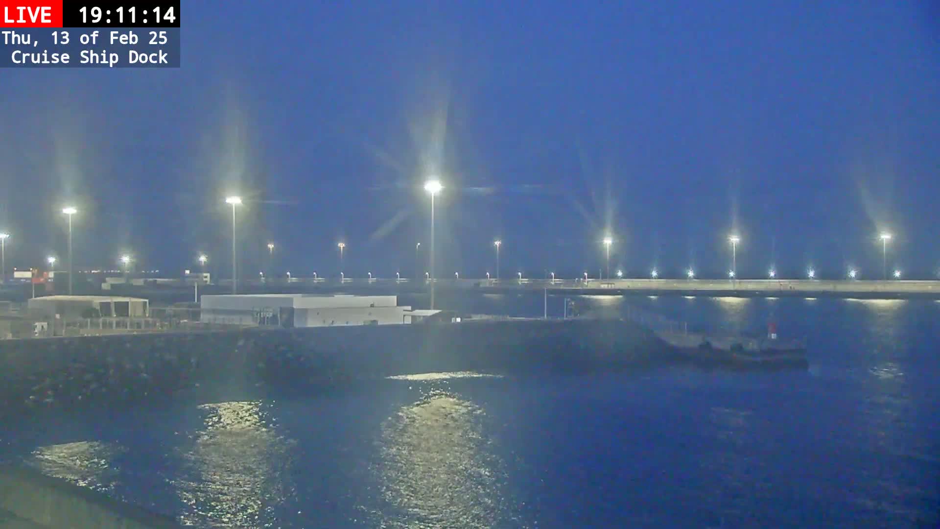 A dark-blue, nighttime view of a cruise ship dock with a small vessel in the water, illuminated by many bright lights.