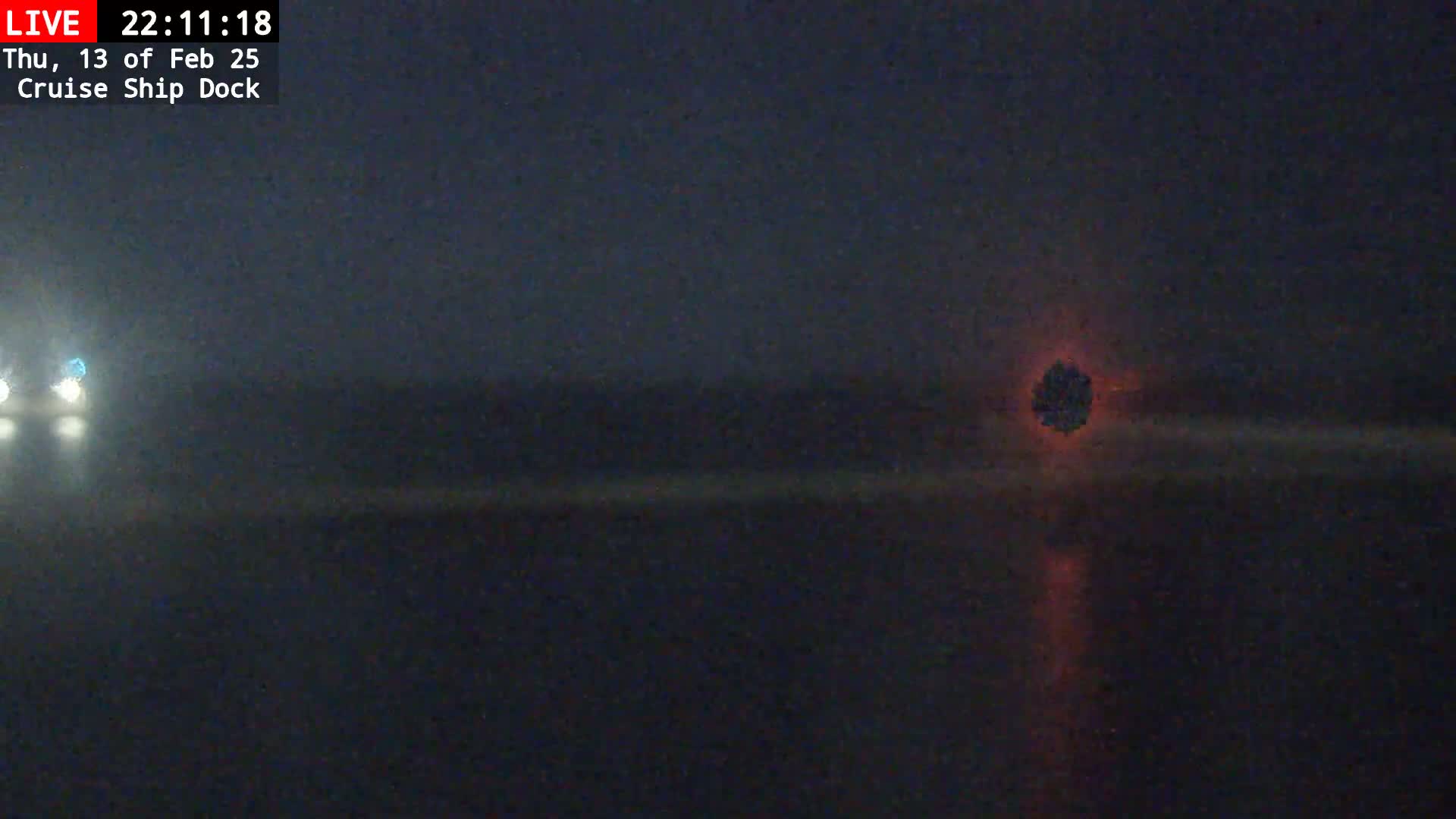 A dark, foggy nighttime scene shows a blurry red light in the distance, with the headlights of a vehicle visible in the foreground.