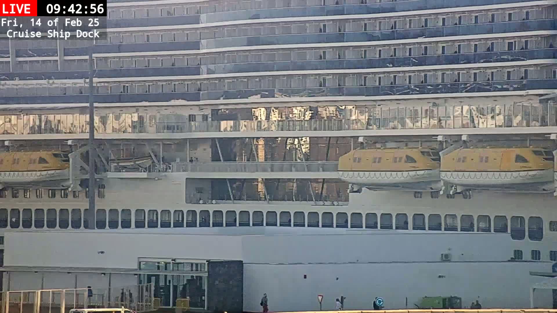 A large cruise ship, showing significant damage to its upper decks, is docked, with two lifeboats visible.