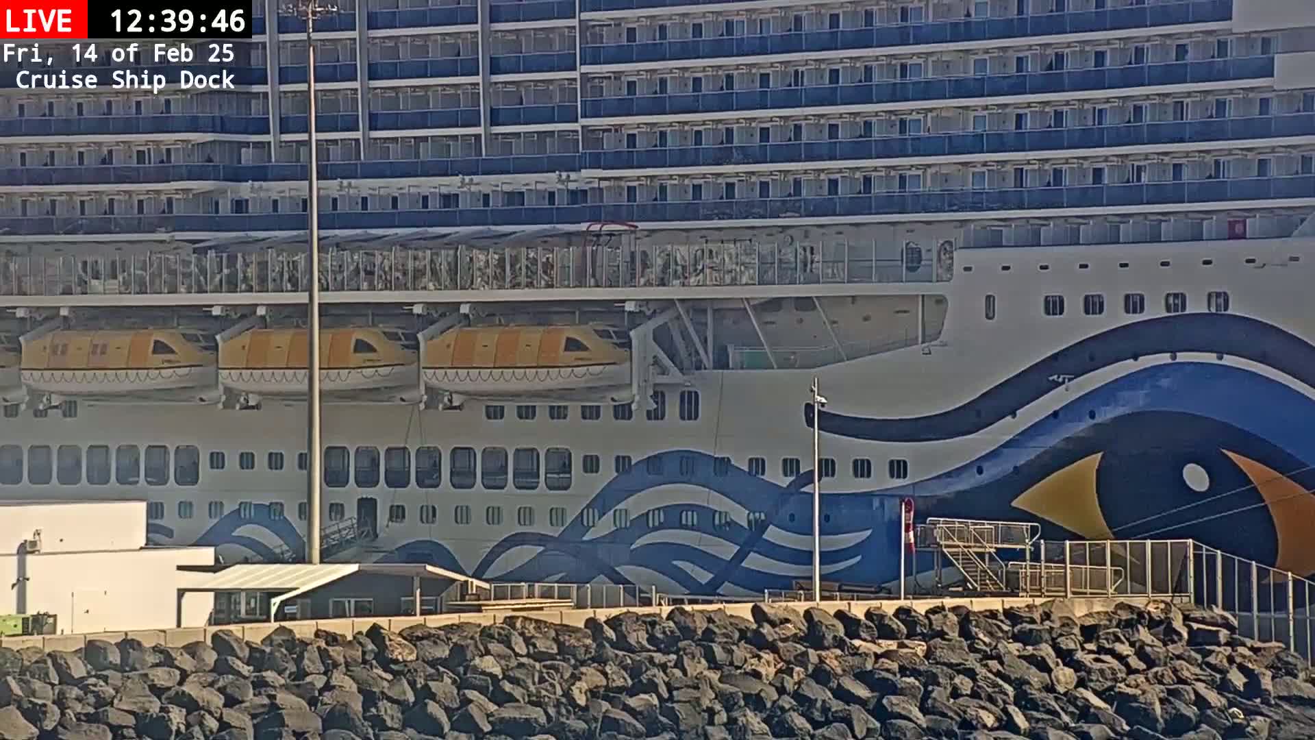 A large cruise ship with yellow lifeboats is docked, and a rocky embankment is in the foreground under a sunny sky.