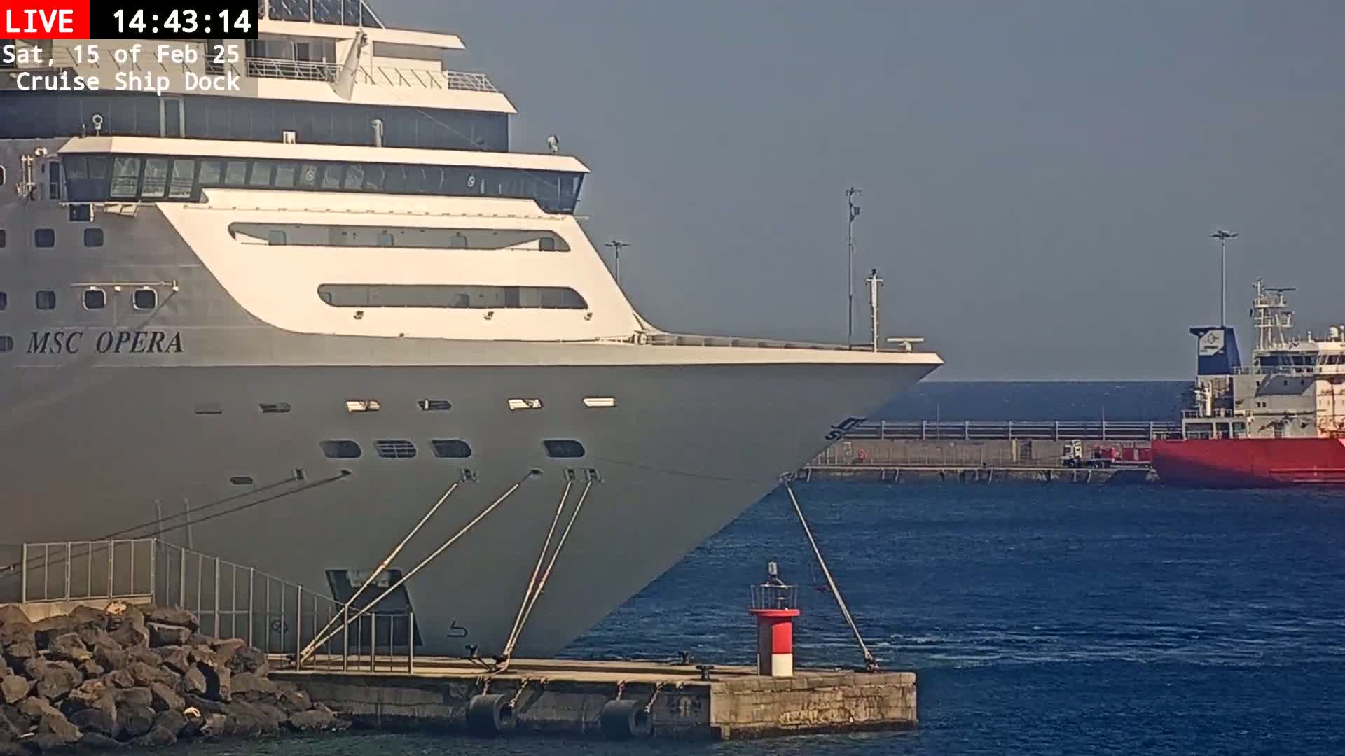 A large cruise ship, the MSC Opera, is docked at a pier on a calm, sunny day, with another smaller, red ship visible in the background.