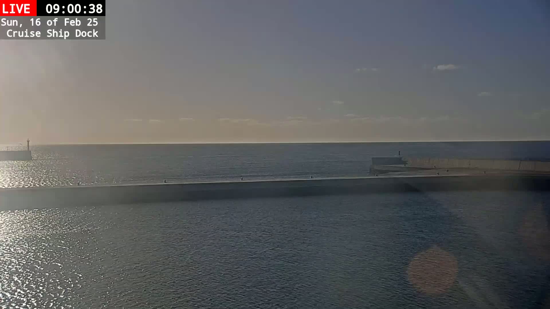 A calm, sunny day is shown overlooking a cruise ship dock with a long, low wall separating the calmer inner water from the open ocean.