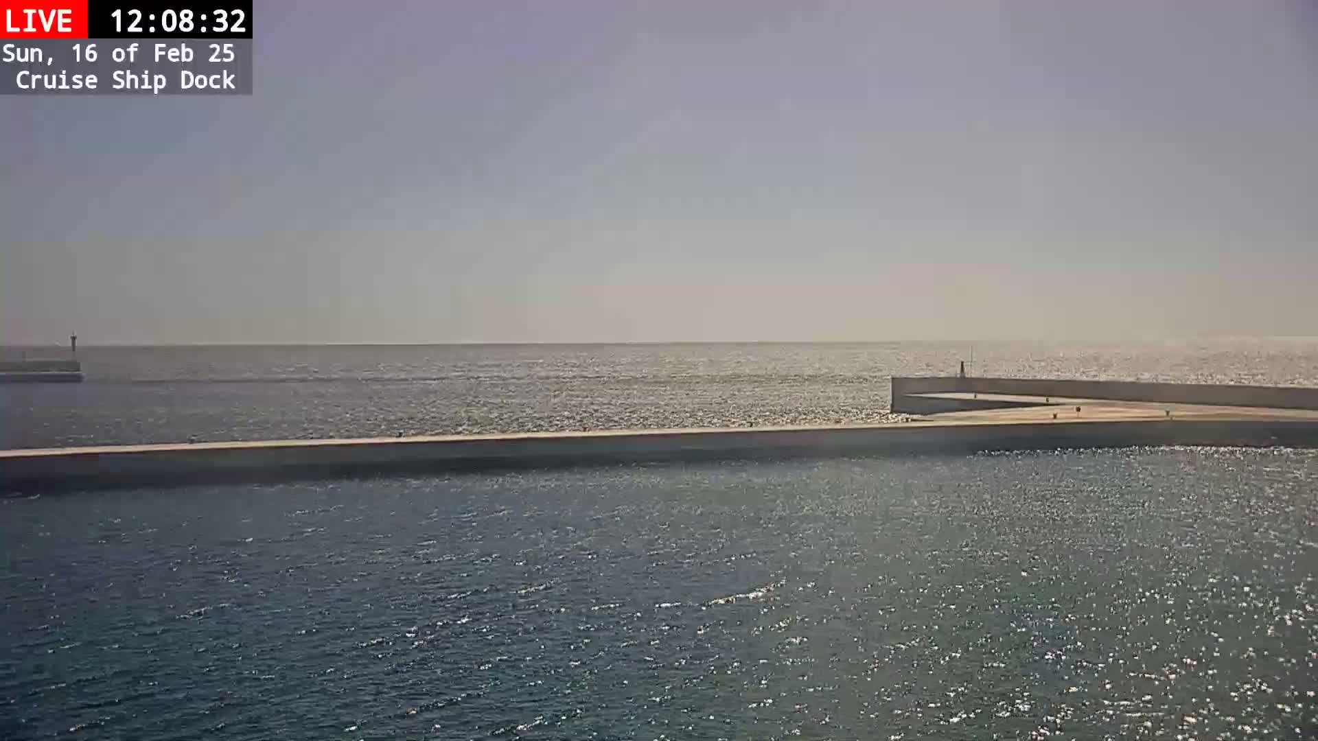 A calm, sunny day is shown at a cruise ship dock with a long, light-colored wall separating a calm body of water from the open ocean.