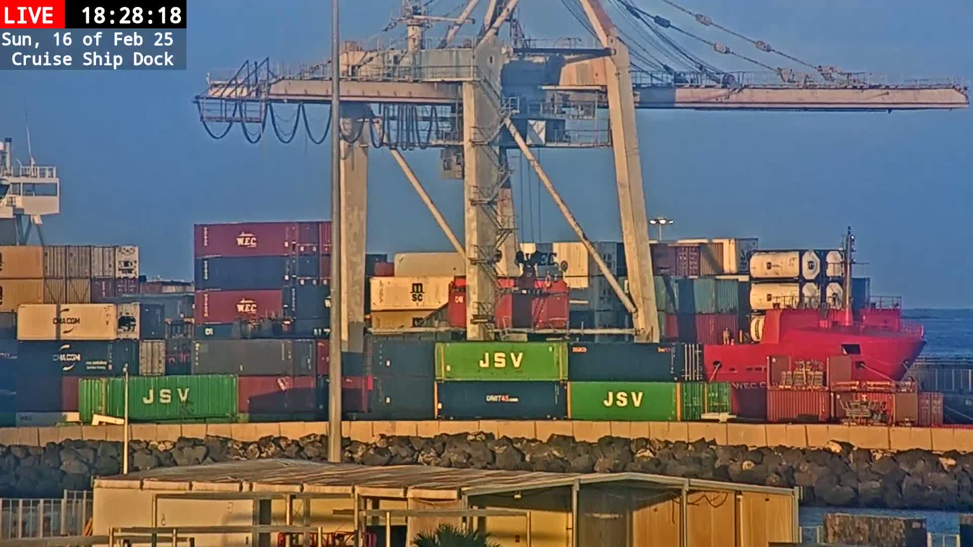 A red cargo ship loaded with shipping containers sits docked at a port under a clear, sunny sky.