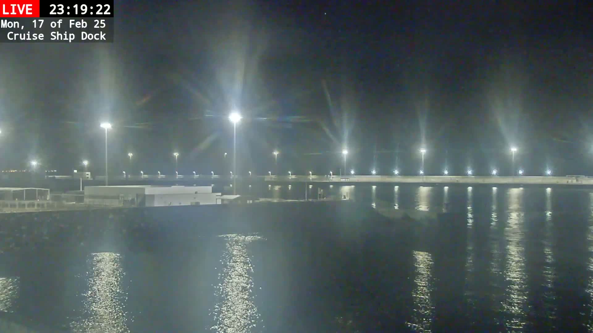A nighttime view of a brightly lit cruise ship dock, with calm water reflecting the lights.