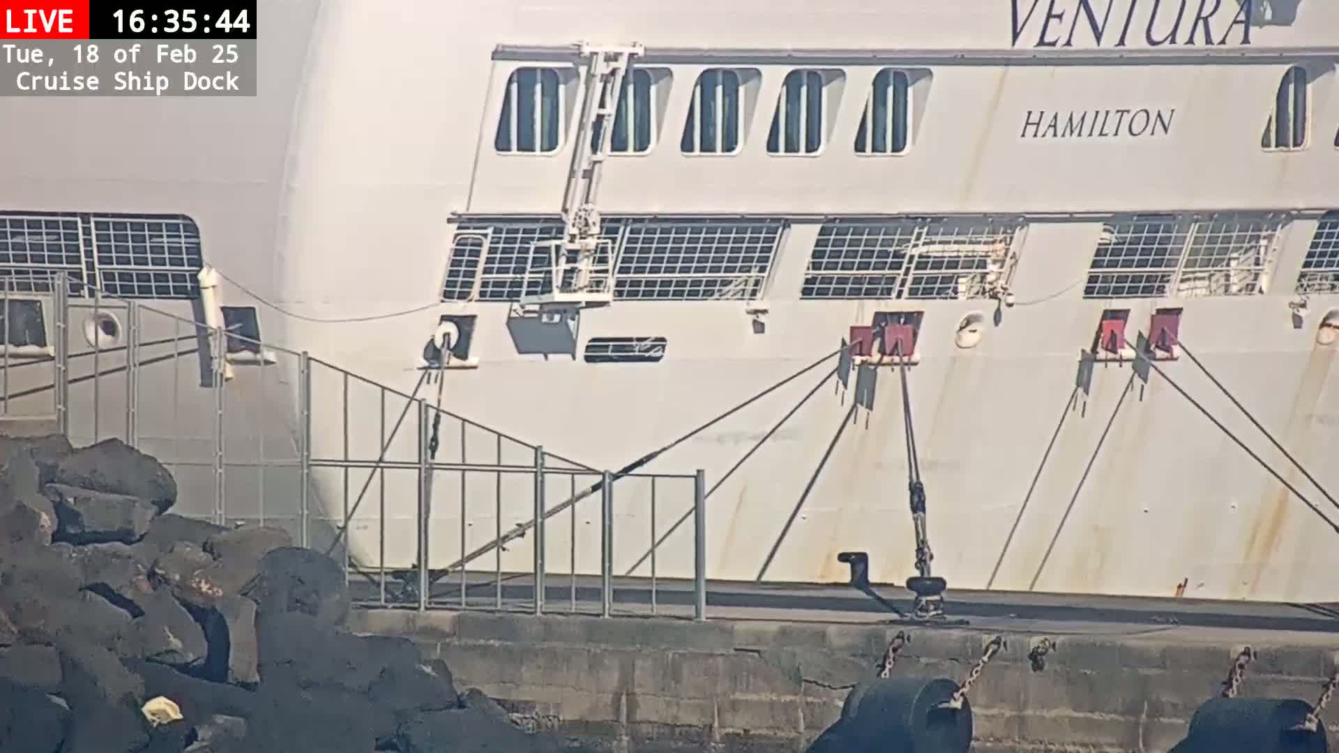 A large, white cruise ship is docked, secured by ropes to a concrete pier with dark rocks along the water's edge, under sunny conditions.