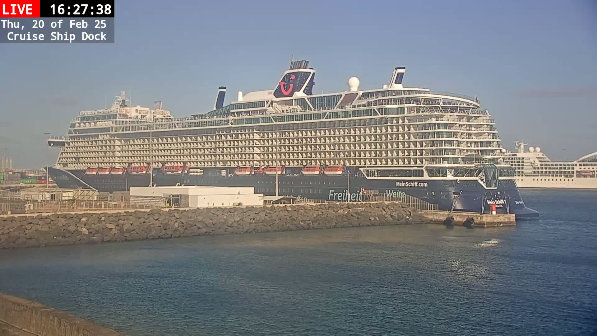 A large cruise ship is docked at a port on a sunny day, with another cruise ship visible in the background.