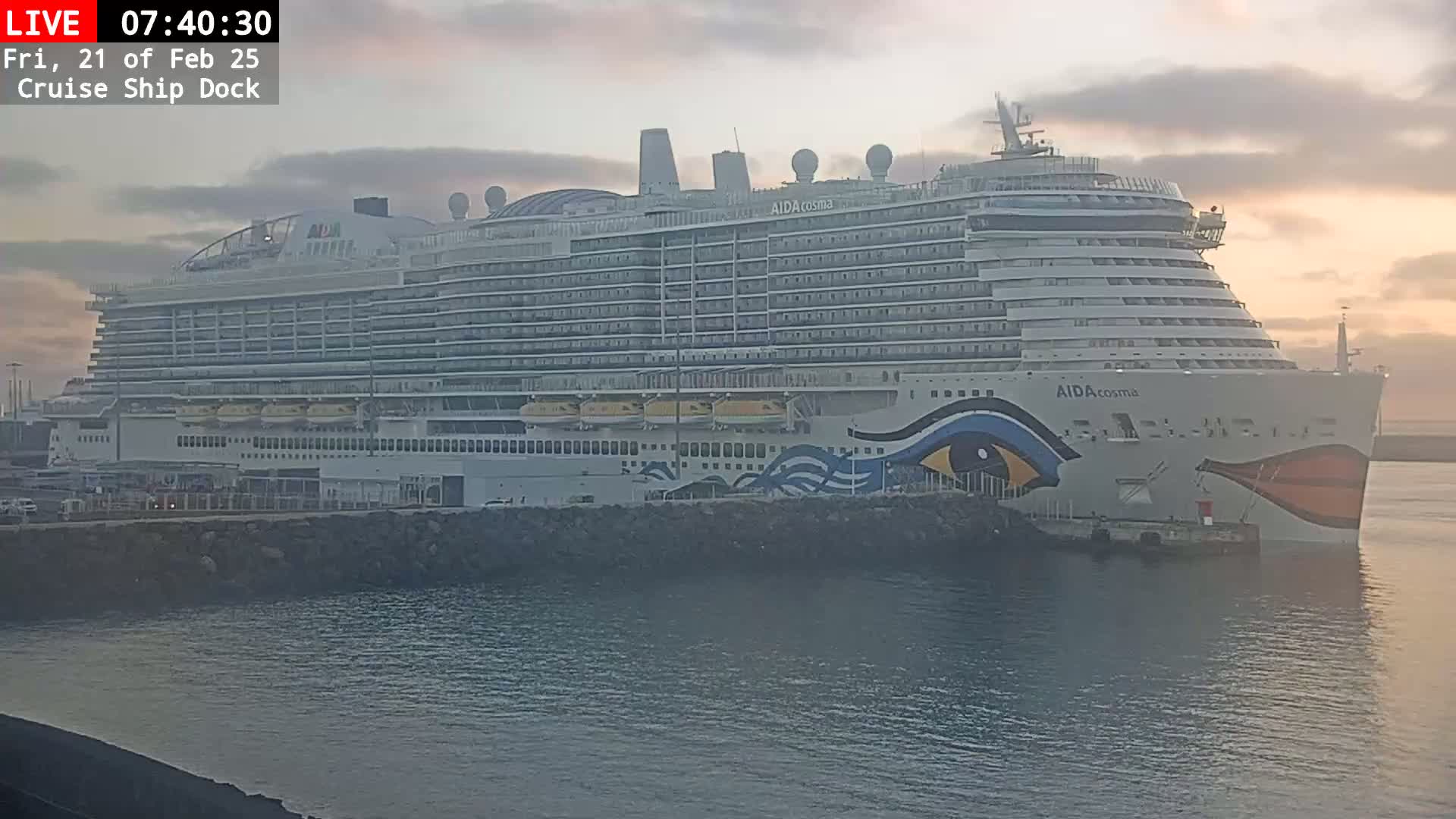 A large cruise ship is docked at a pier during a sunrise with a mostly cloudy sky.
